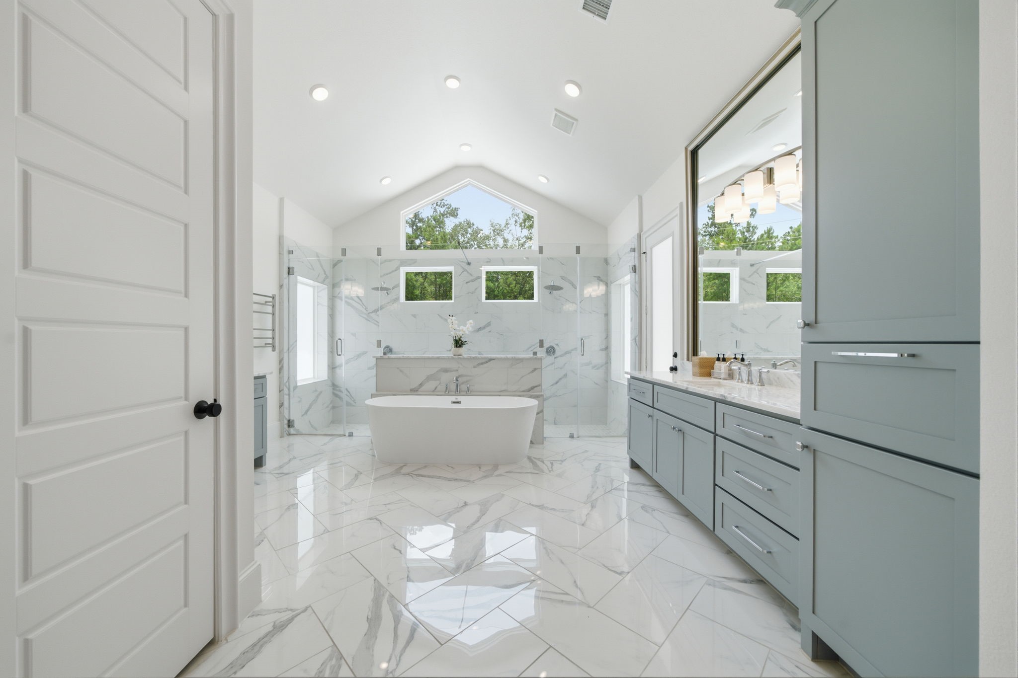 Luxurious bathroom featuring a freestanding tub, sleek marble flooring, and dual vanities. The vaulted ceiling and large windows provide abundant natural light.