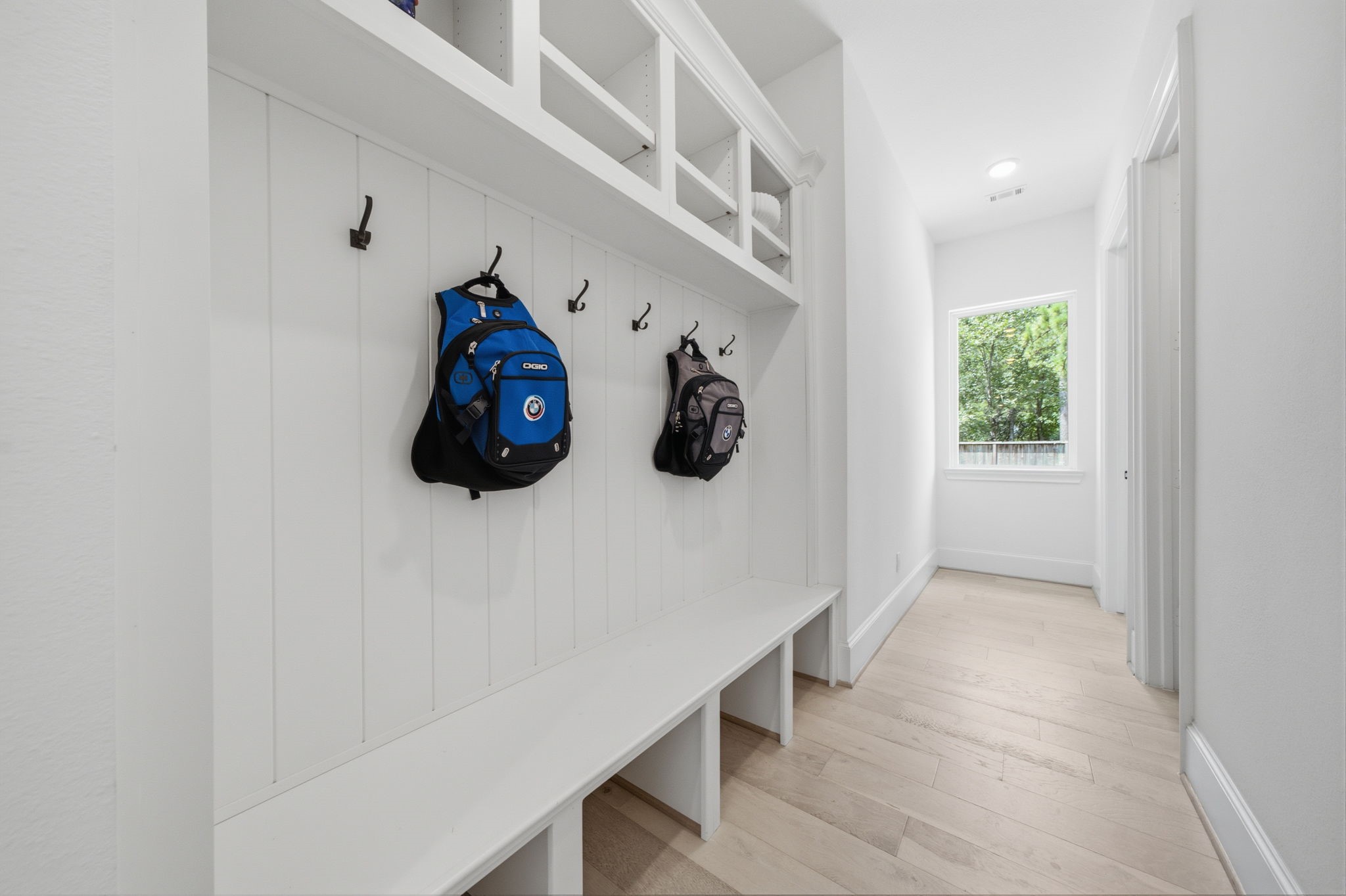 Spacious mudroom featuring built-in storage with hooks and cubbies, complemented by sleek white paneling and light wood flooring, offering a practical and stylish entry solution.