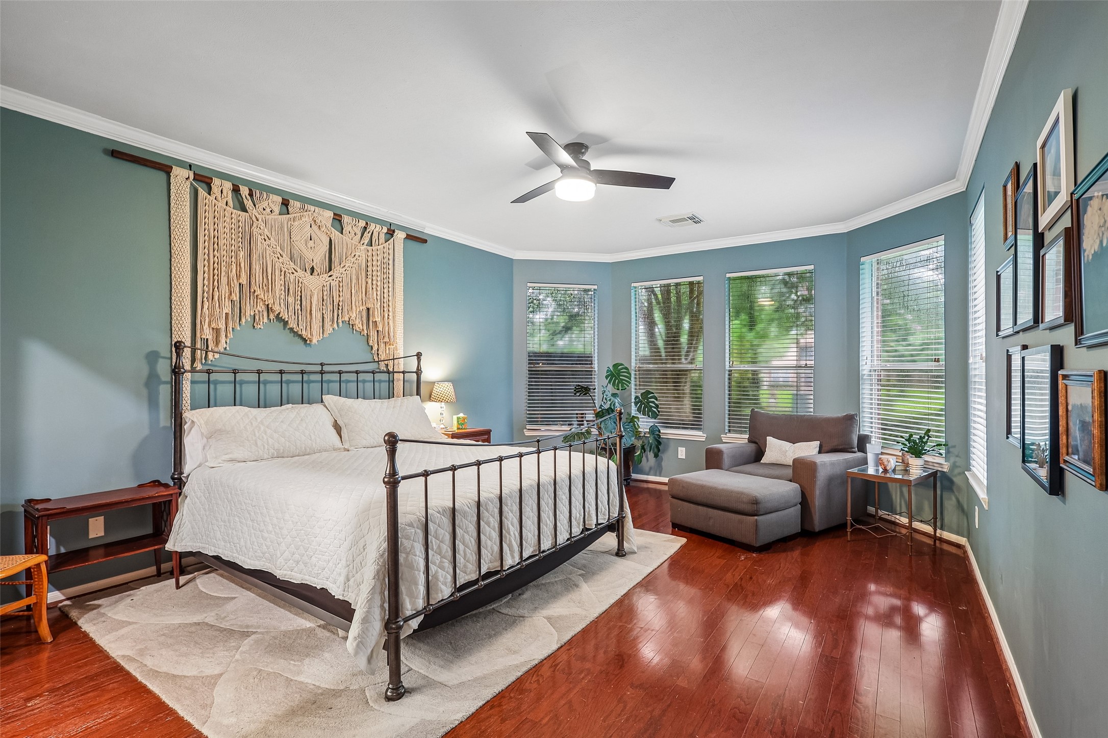 This spacious bedroom features hardwood floors, soft blue walls, and ample natural light from multiple large windows. It includes a stylish metal bedframe, cozy seating area, and decorative wall hangings, creating a warm and inviting atmosphere.