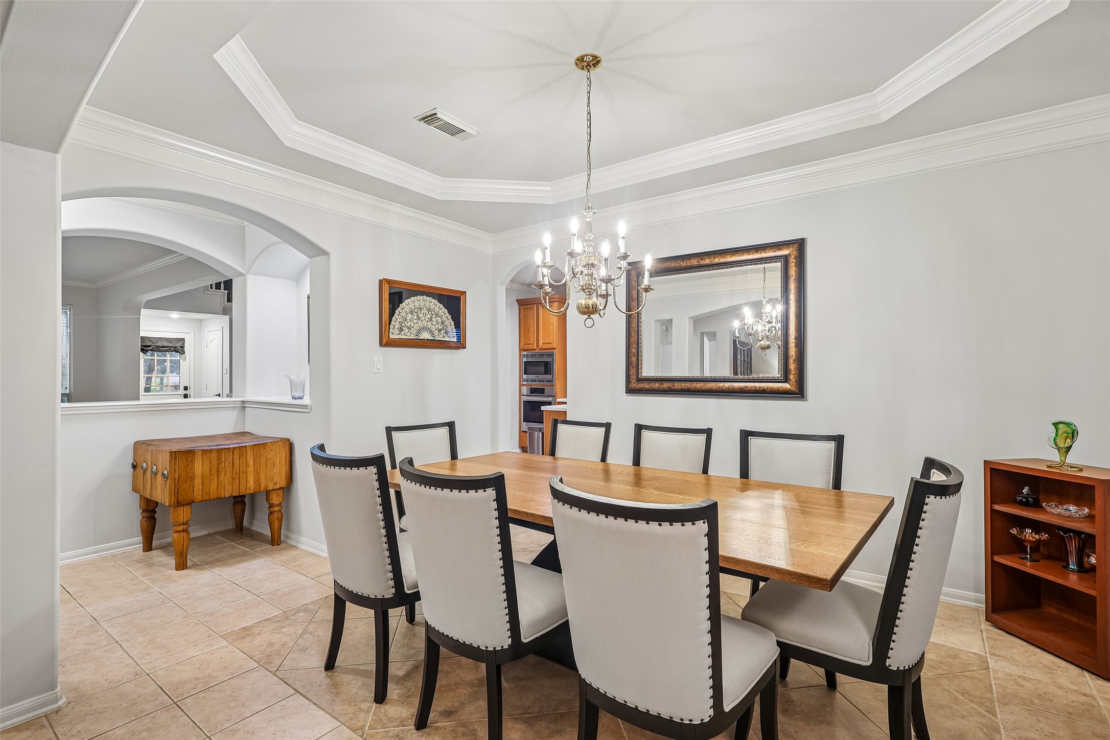 This elegant dining room features a large wooden table with upholstered chairs, a classic chandelier, and a tray ceiling. The space is bright and open, with arched doorways and tile flooring, offering a welcoming atmosphere for gatherings.