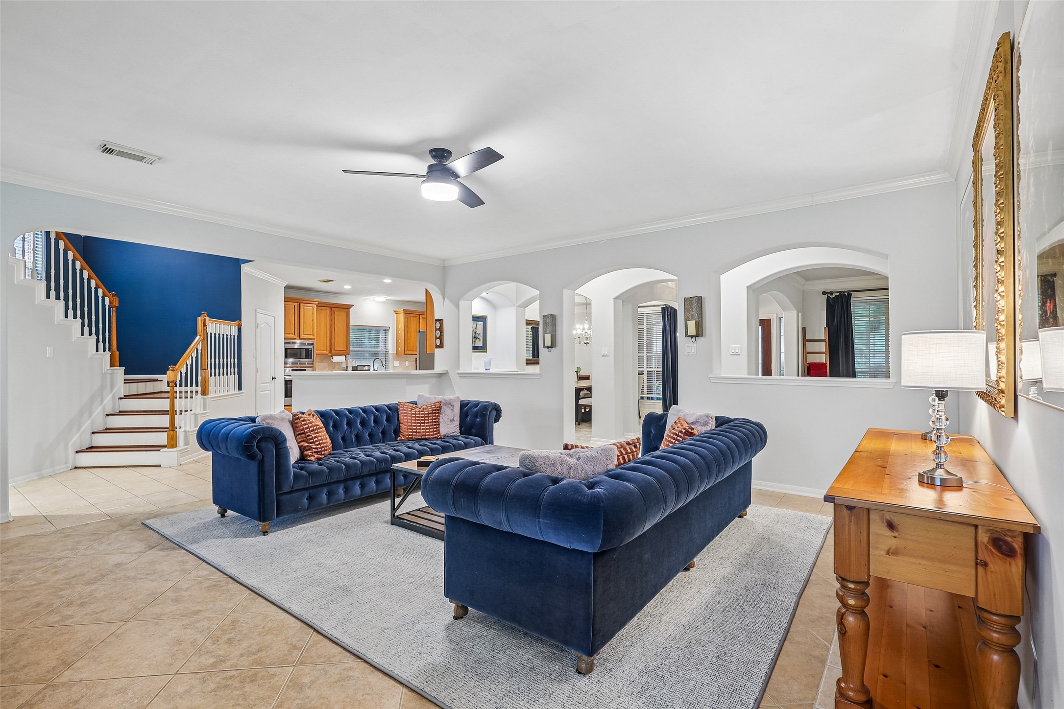 This inviting living room features elegant navy sofas, a modern ceiling fan, and a cozy atmosphere. The open layout connects seamlessly to a spacious kitchen with warm wooden cabinetry, while arched doorways add a touch of sophistication. A stairway with white and wooden accents leads to the upper level, enhancing the home's charm. This inviting living room features elegant navy sofas, a modern ceiling fan, and a cozy atmosphere. The open layout connects seamlessly to a spacious kitchen with warm wooden cabinetry, while arched doorways add a touch of sophistication. A stairway with white and wooden accents leads to the upper level, enhancing the home's charm.