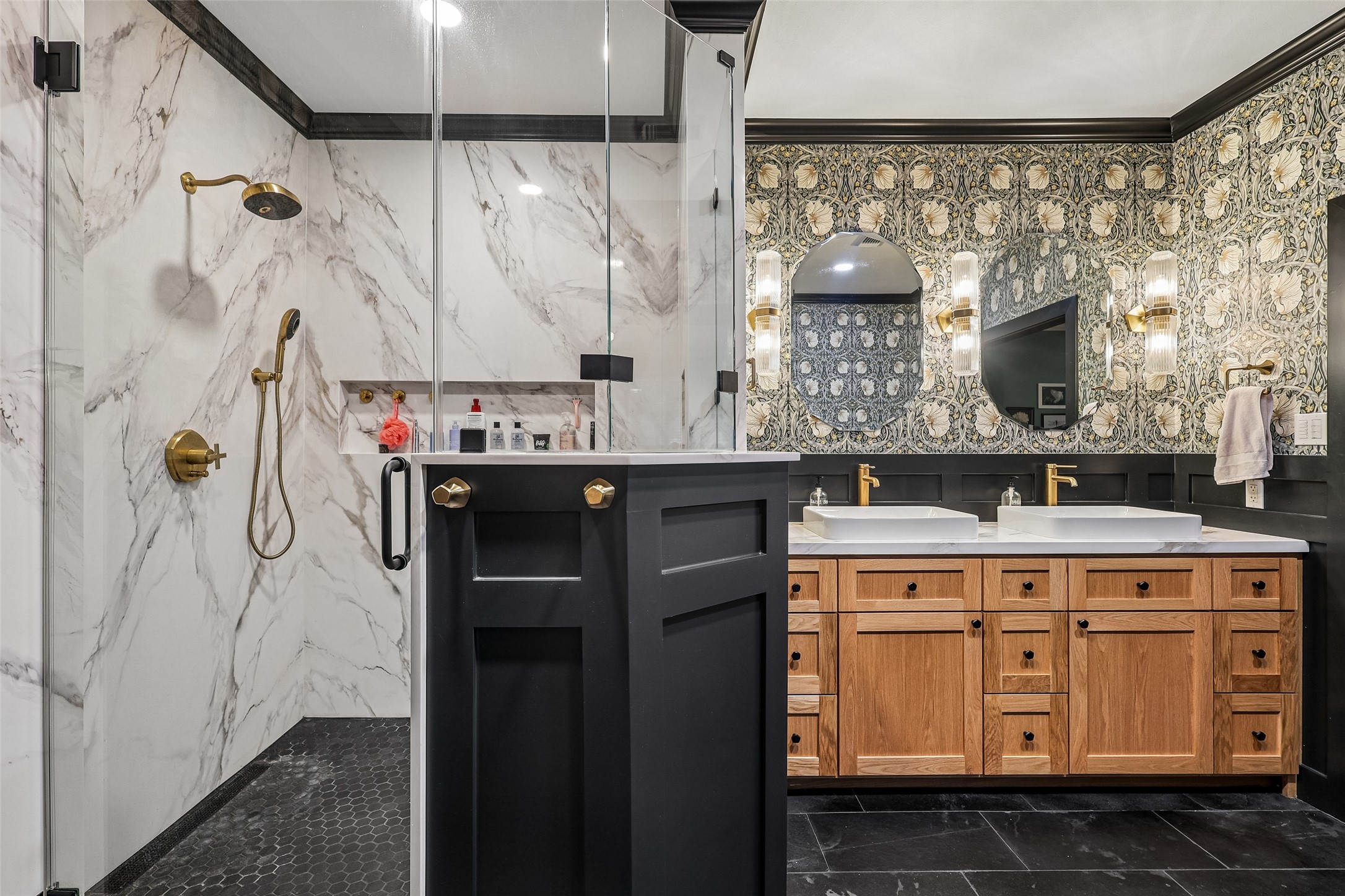 This bathroom features a luxurious design with a spacious glass-enclosed shower with marble walls, dual sinks with elegant wooden cabinetry, and stylish patterned wallpaper. Gold accents and modern lighting complete the sophisticated look.