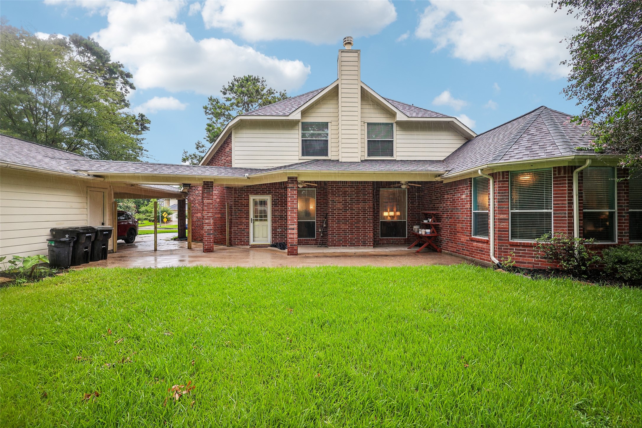 This photo shows a charming two-story brick home with a spacious, well-maintained backyard. It features a covered patio area and a detached garage connected by a carport. The exterior is accented with large windows and a chimney, offering a cozy and inviting atmosphere.