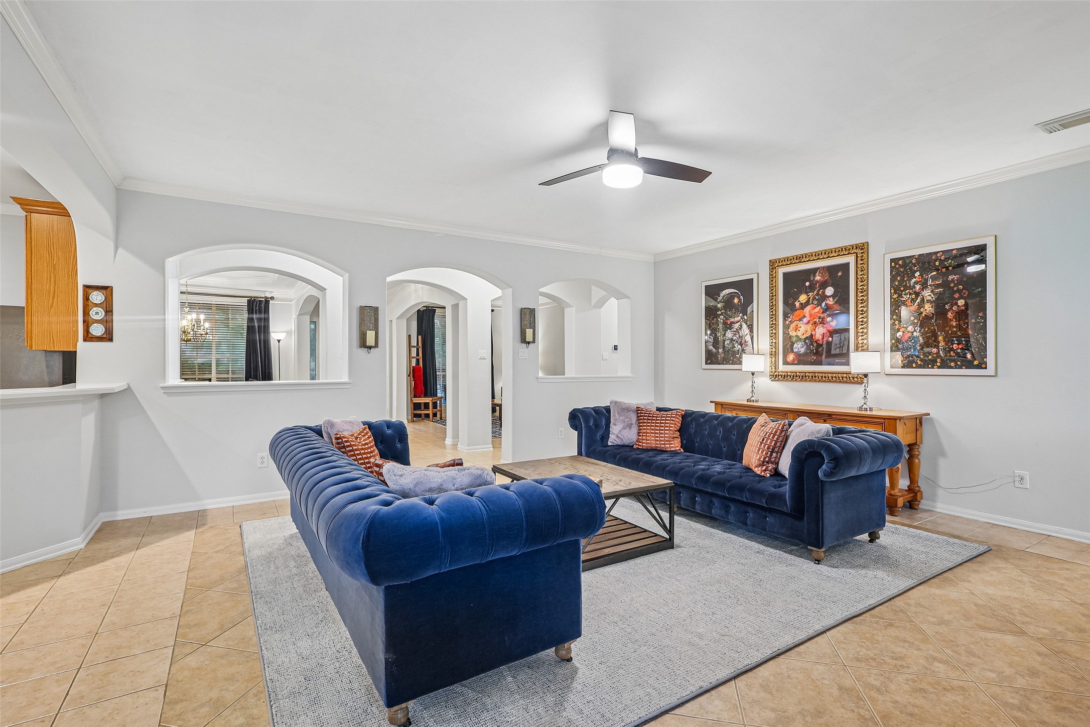 Spacious living room with elegant blue sofas and modern decor. Features tiled flooring, a ceiling fan, and stylish wall art. Arched doorways create an open, inviting atmosphere with plenty of natural light. Spacious living room with elegant blue sofas and modern decor. Features tiled flooring, a ceiling fan, and stylish wall art. Arched doorways create an open, inviting atmosphere with plenty of natural light.