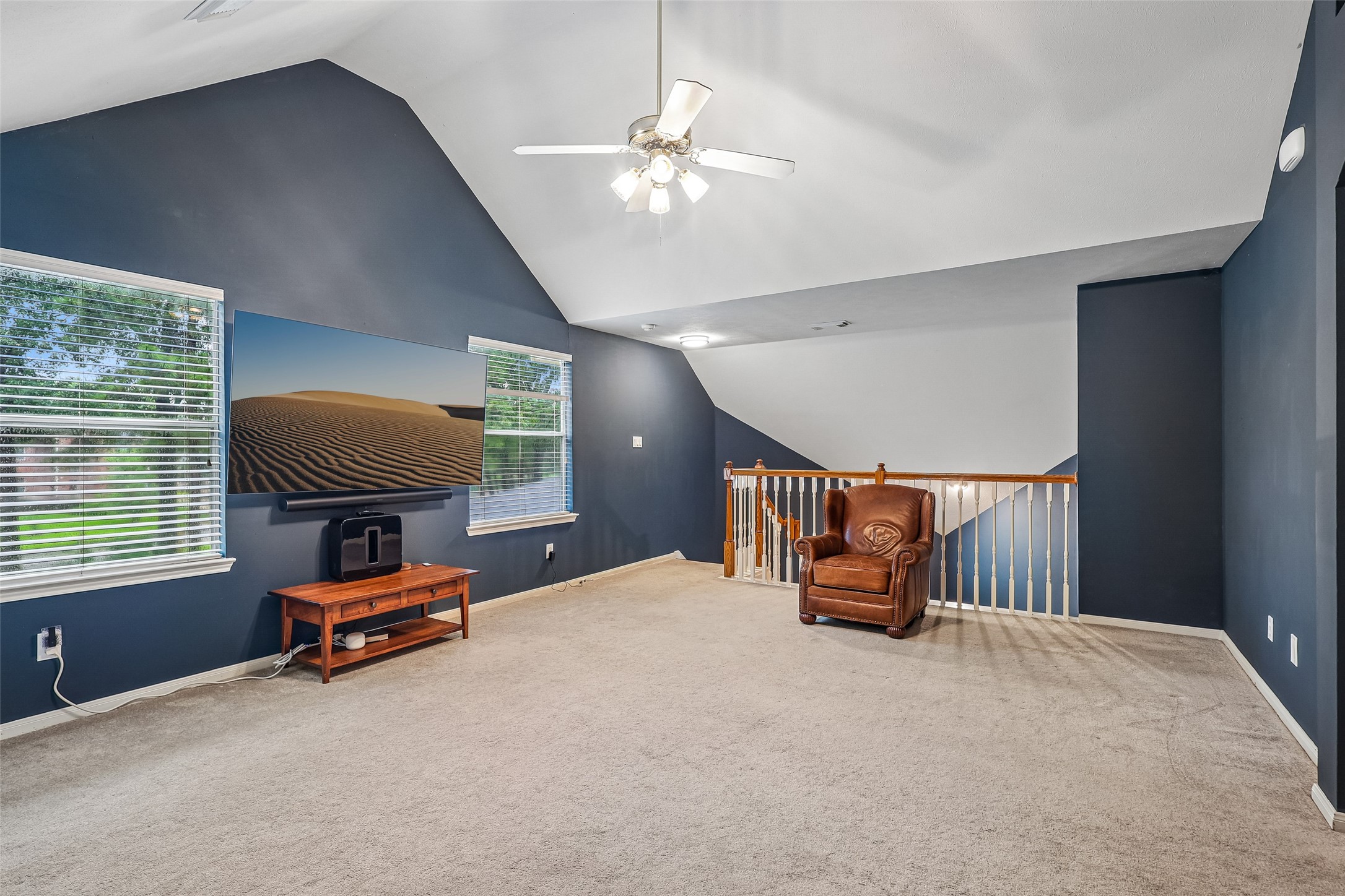 Bright and spacious upstairs living area with vaulted ceilings, featuring a ceiling fan, cozy seating, and a large TV. Two windows provide ample natural light, enhancing the room's inviting atmosphere.