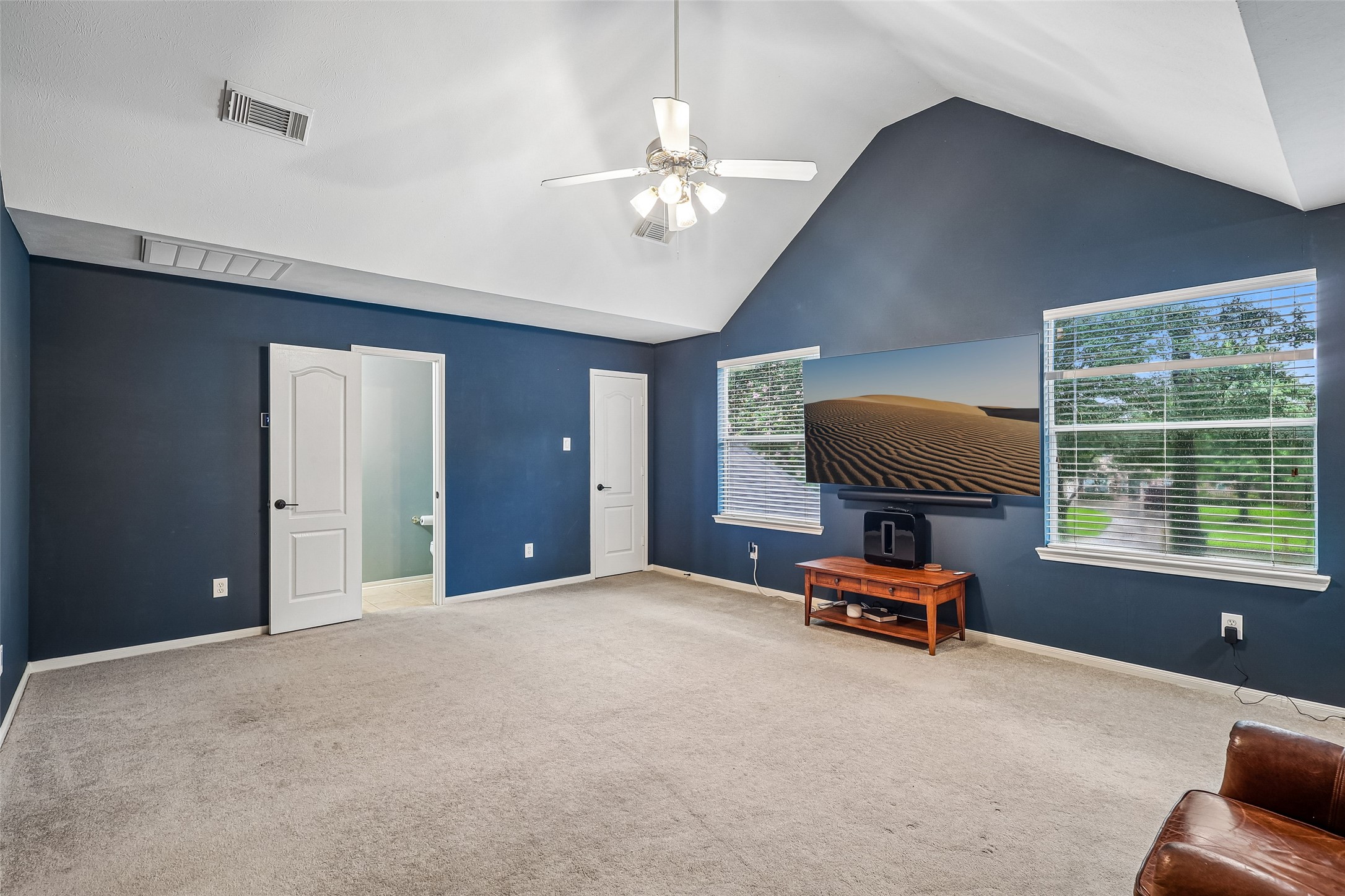 Spacious room with vaulted ceilings, large windows, and a ceiling fan. Features carpeted flooring, blue accent walls, and a mounted TV. Ideal for a cozy living area or media room.