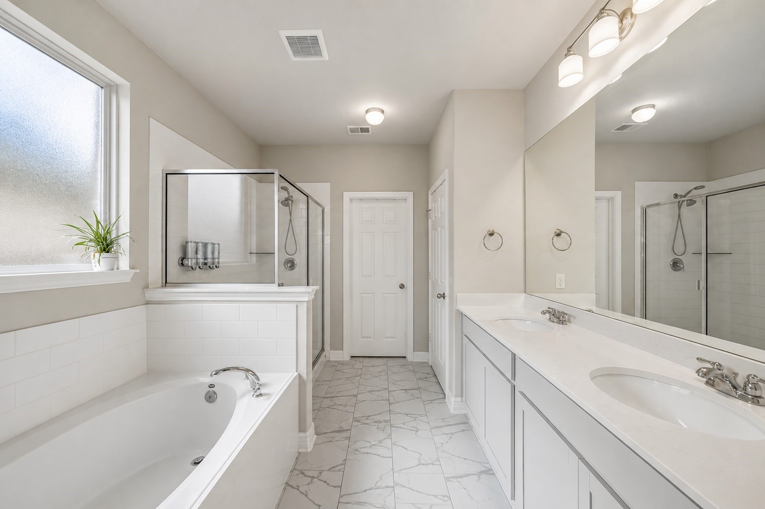 This spacious primary bathroom features a bright, neutral design with a long dual-sink vanity, expansive mirror, and generous counter and cabinet space. A large soaking tub and separate glass-enclosed shower create a relaxing, spa-inspired setup, while marble-look tile flooring and abundant natural light give the space a clean, polished feel.