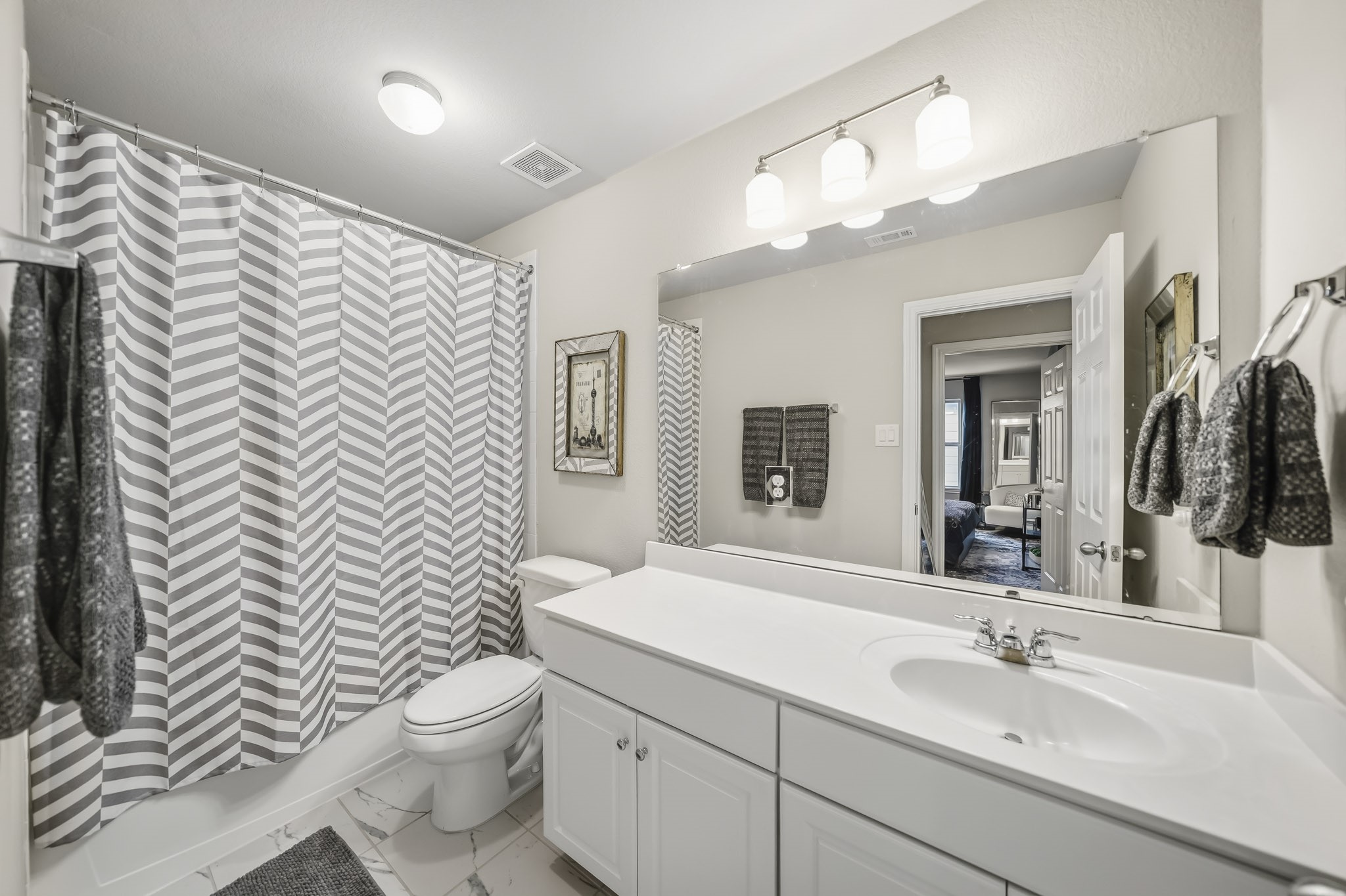 This bright secondary bathroom features a clean, neutral design with a spacious vanity, generous countertop space, and a large wall-to-wall mirror. A tub and shower combination adds everyday functionality, while updated tile flooring and ample lighting create a fresh, inviting feel.
