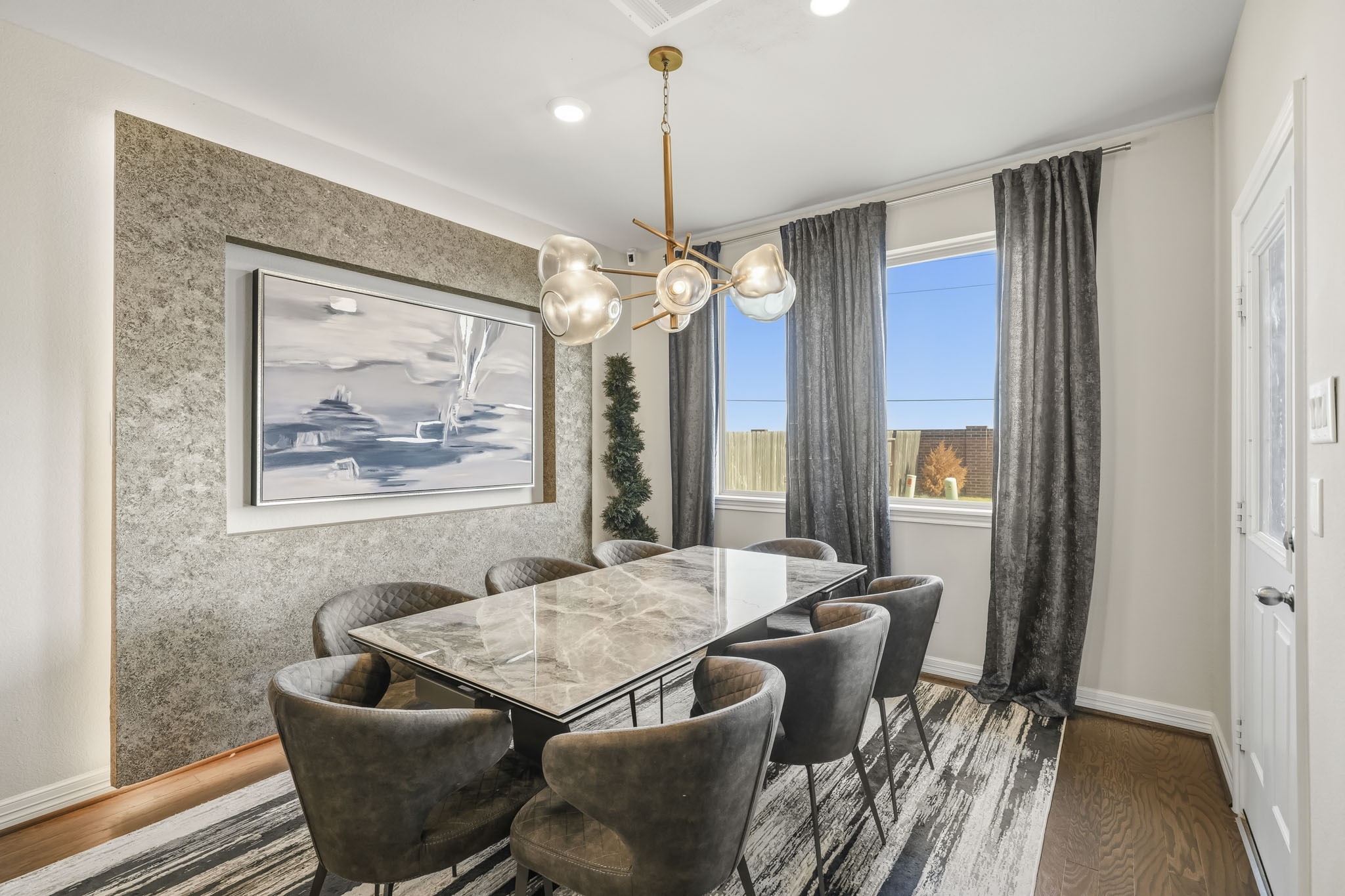 This elegant dining area features warm wood flooring, a sophisticated statement chandelier, and a striking textured accent wall that adds depth and character. A large window fills the space with natural light, while the generous layout comfortably accommodates a large dining table, making it ideal for everyday meals and entertaining guests.