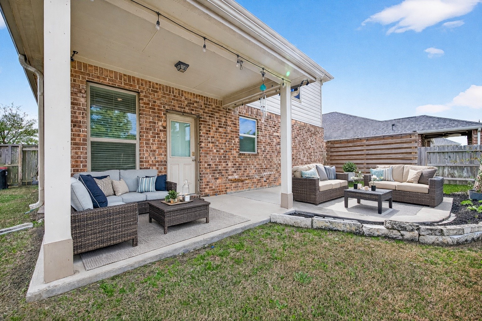 This photo showcases a cozy backyard patio with comfortable outdoor seating on a concrete slab, set against a brick exterior. The area is partially covered, featuring string lights and a small garden space bordered by stone edging, perfect for outdoor relaxation and entertaining. (enhanced photo) This photo showcases a cozy backyard patio with comfortable outdoor seating on a concrete slab, set against a brick exterior. The area is partially covered, featuring string lights and a small garden space bordered by stone edging, perfect for outdoor relaxation and entertaining. (enhanced photo)
