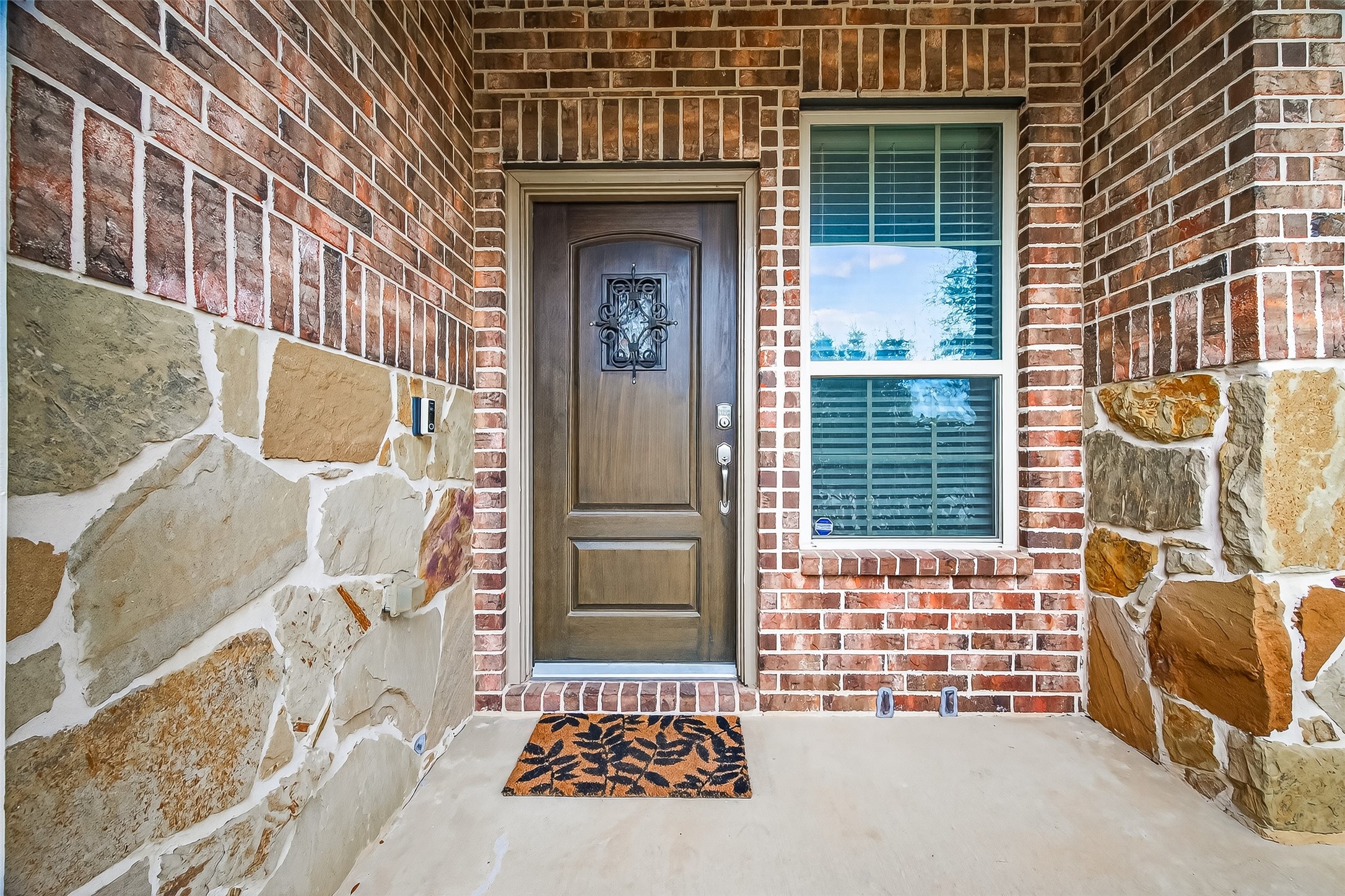 This photo showcases a welcoming entrance with a sturdy wooden door featuring decorative ironwork. It's framed by brick and stone walls, adding a rustic charm. A large window with shutters allows natural light inside, and a doormat adds a cozy touch.