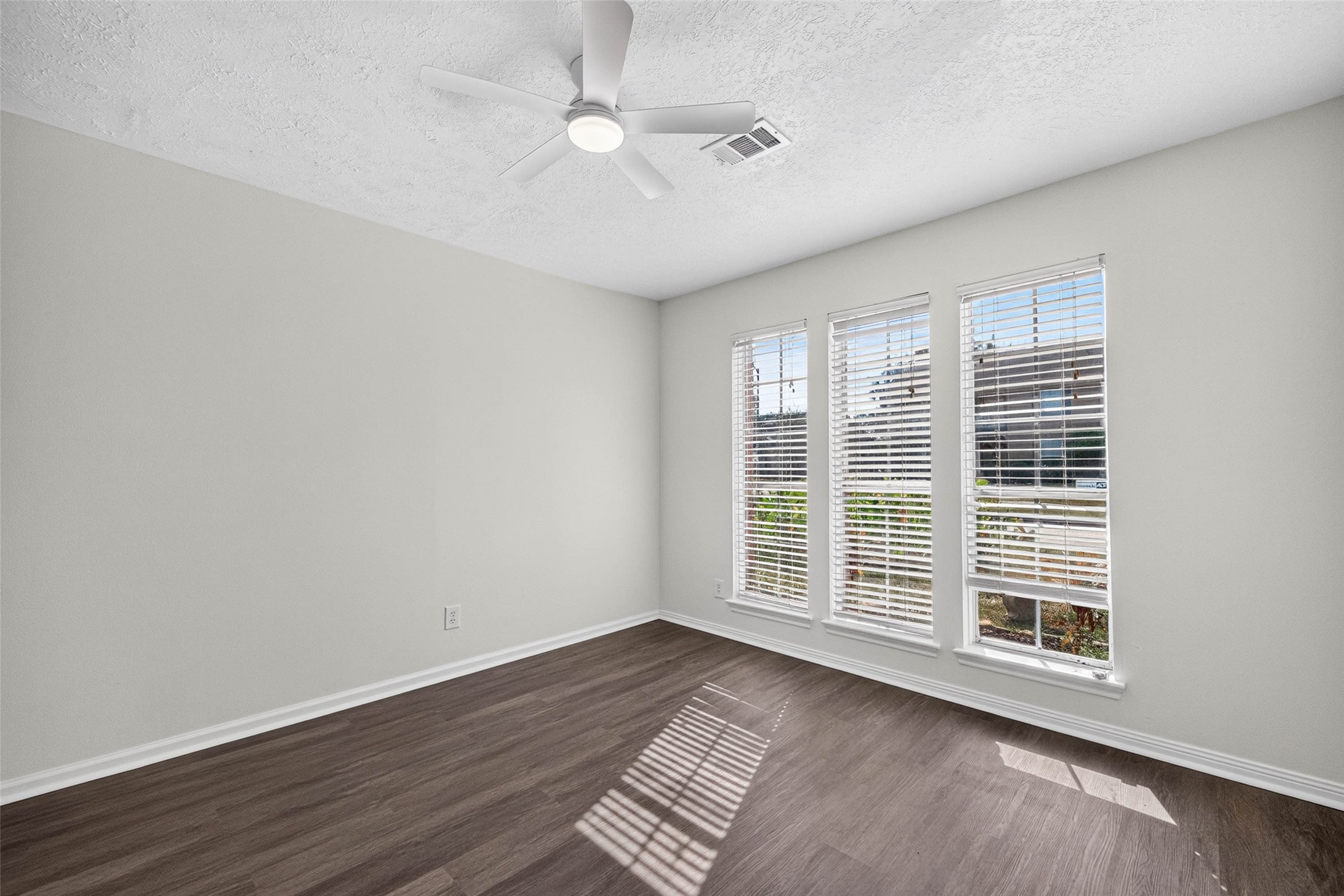 Bedroom with wood-look flooring and three tall windows with blinds, brightened by a ceiling fan with light.