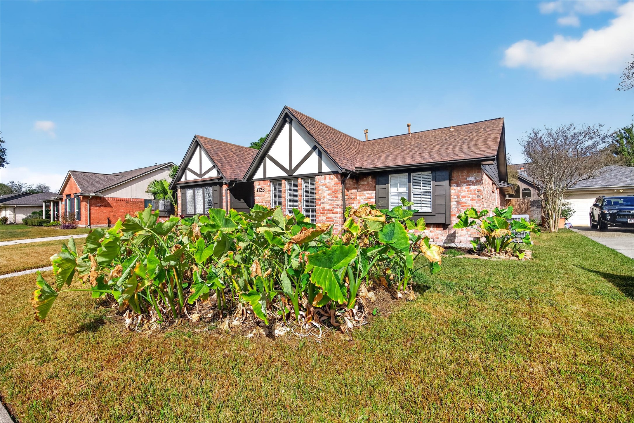 Angled front view across the lawn to the brick facade and white gables with dark timber trim, with leafy plants in the front bed and the driveway at the side.