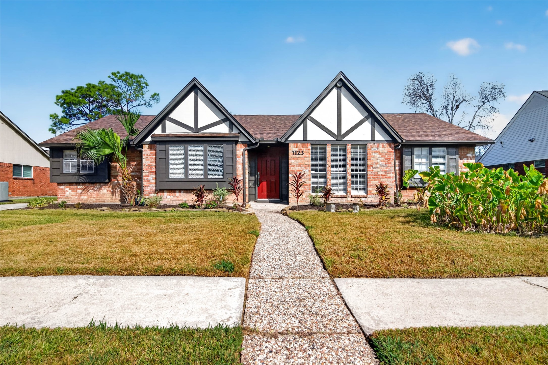 Front view of the single-story ranch, with brick walls, white gables with dark timber trim, a brown shingle roof, and a red front door reached by a pebbled concrete walk. Front view of the single-story ranch, with brick walls, white gables with dark timber trim, a brown shingle roof, and a red front door reached by a pebbled concrete walk.