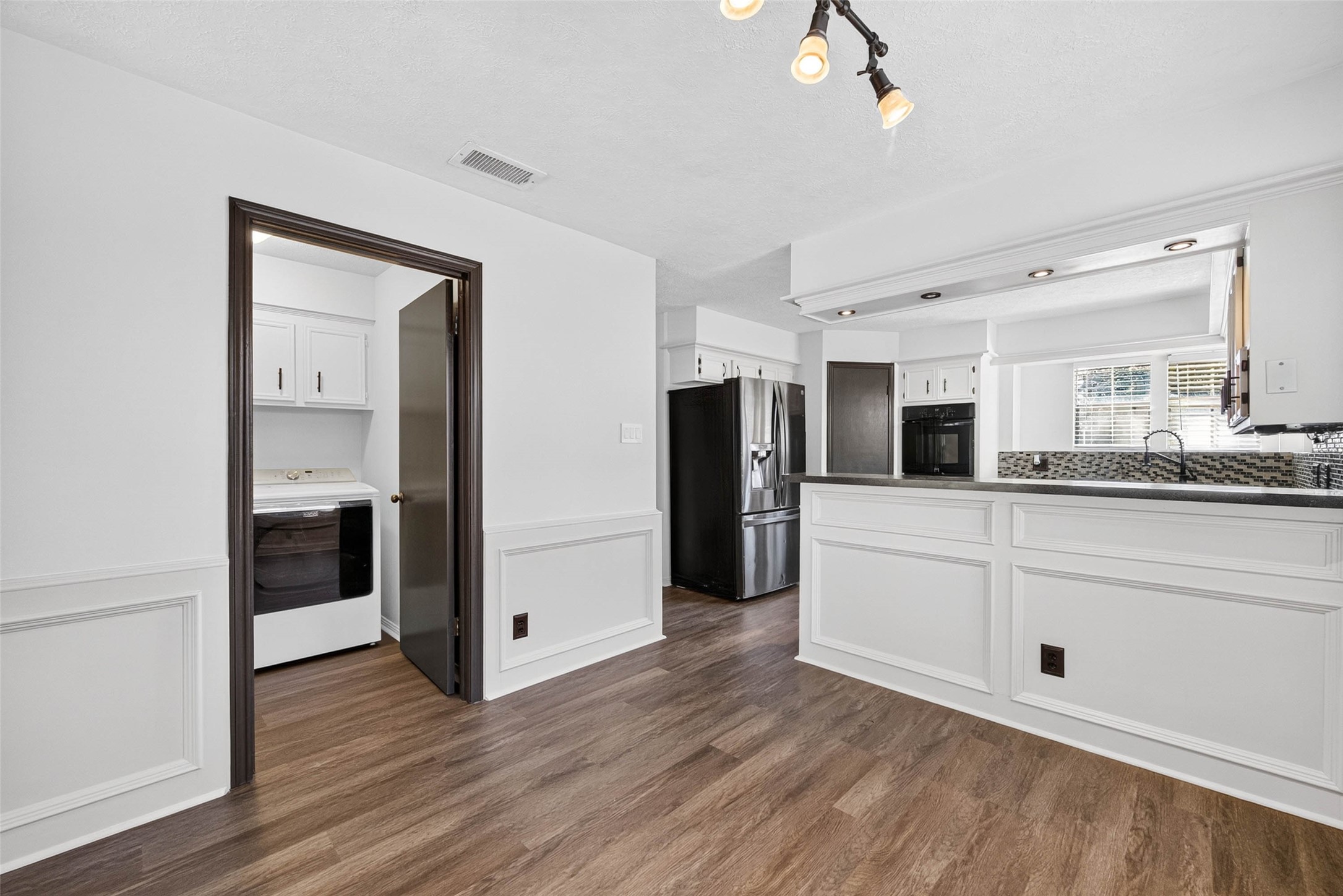 View from the dining area toward the kitchen peninsula, with a stainless refrigerator, black double oven, and a doorway opening to the laundry room.