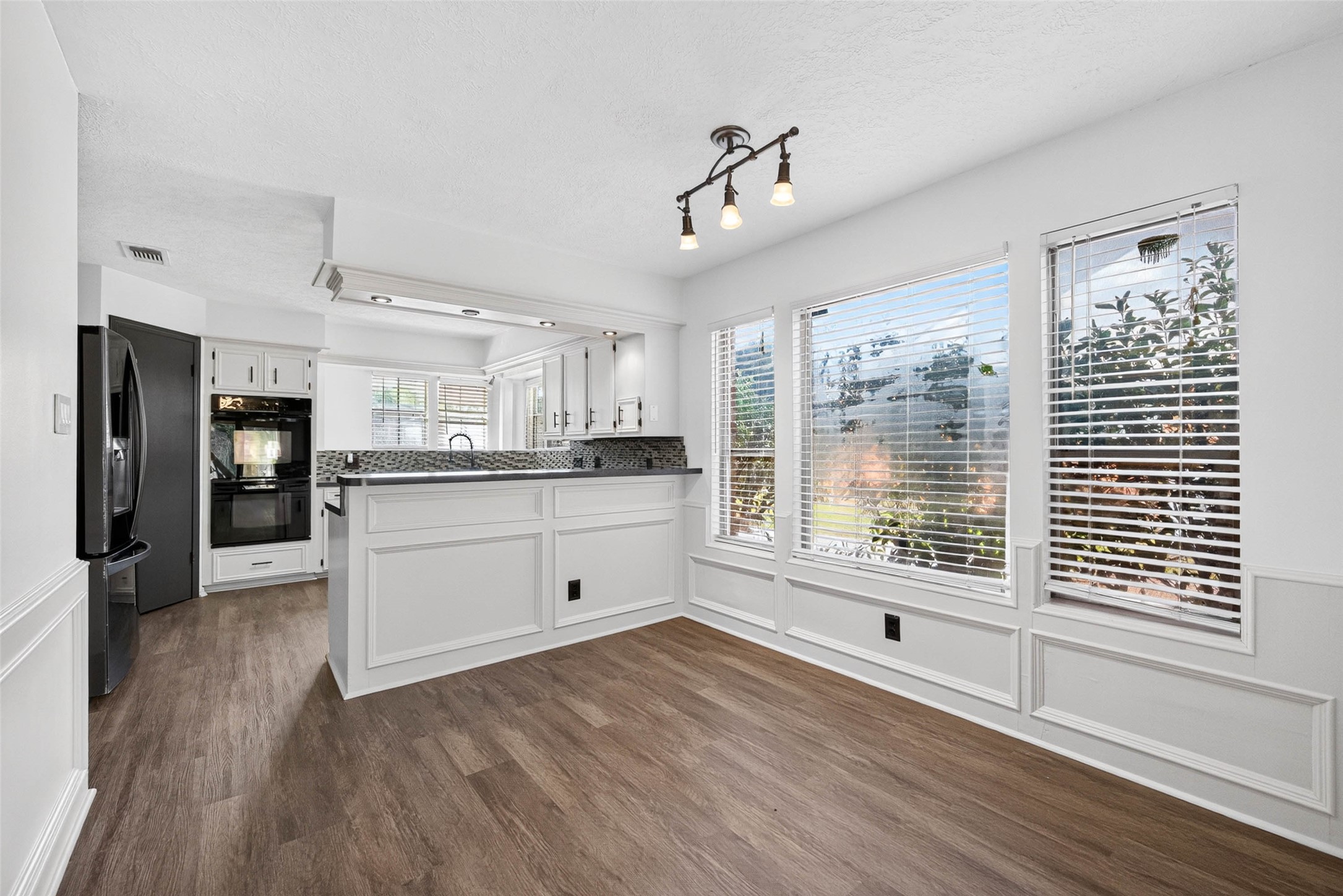 This dining and kitchen space features a paneled peninsula, a mosaic tile backsplash, and a built-in black double wall oven. Large windows with blinds let in natural light, framing views over the wood-look flooring.