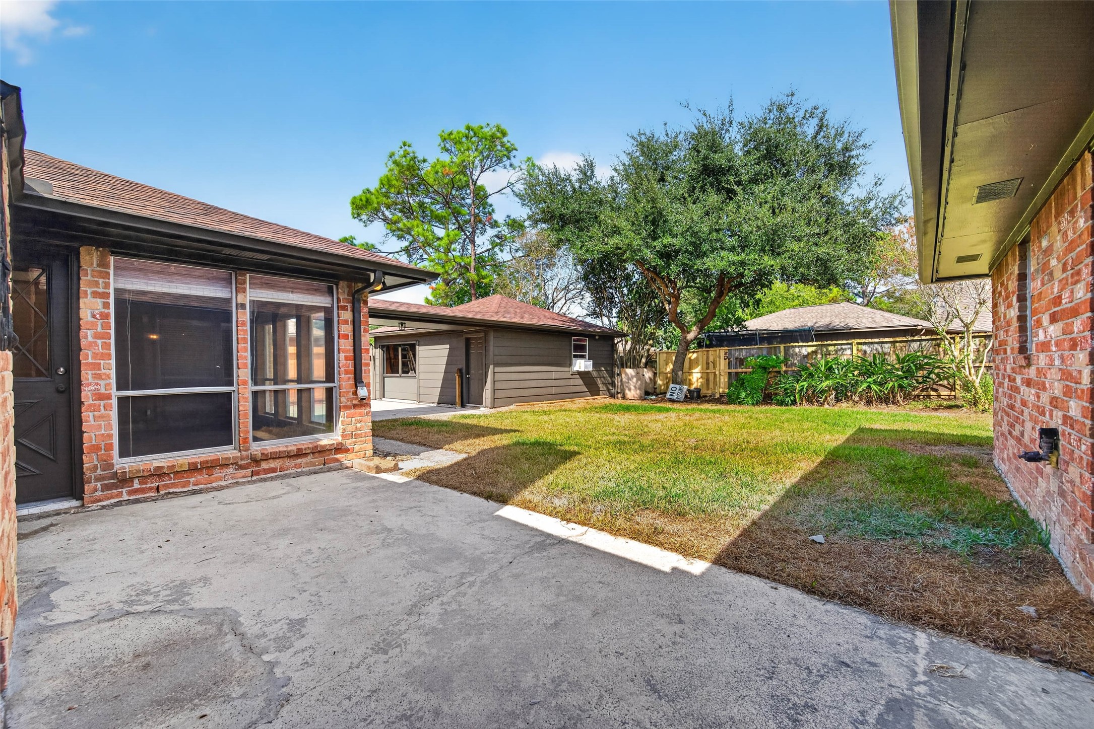 Backyard view with the brick house wall, large windows, lawn, mature trees, and wooden fencing.