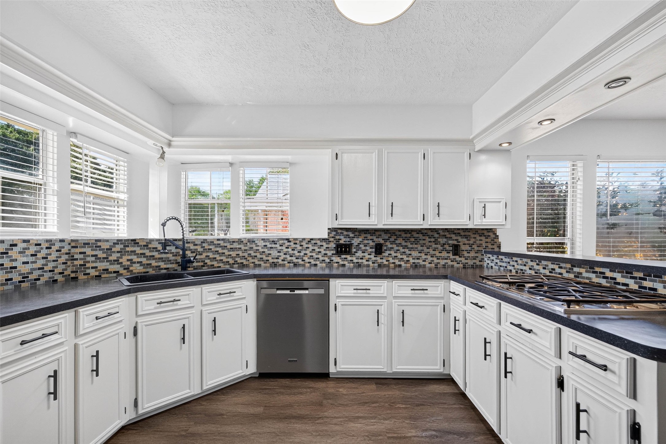 Straight-on kitchen view with cabinets, mosaic tile backsplash, double sink beneath windows, stainless dishwasher, and gas cooktop on the side peninsula.