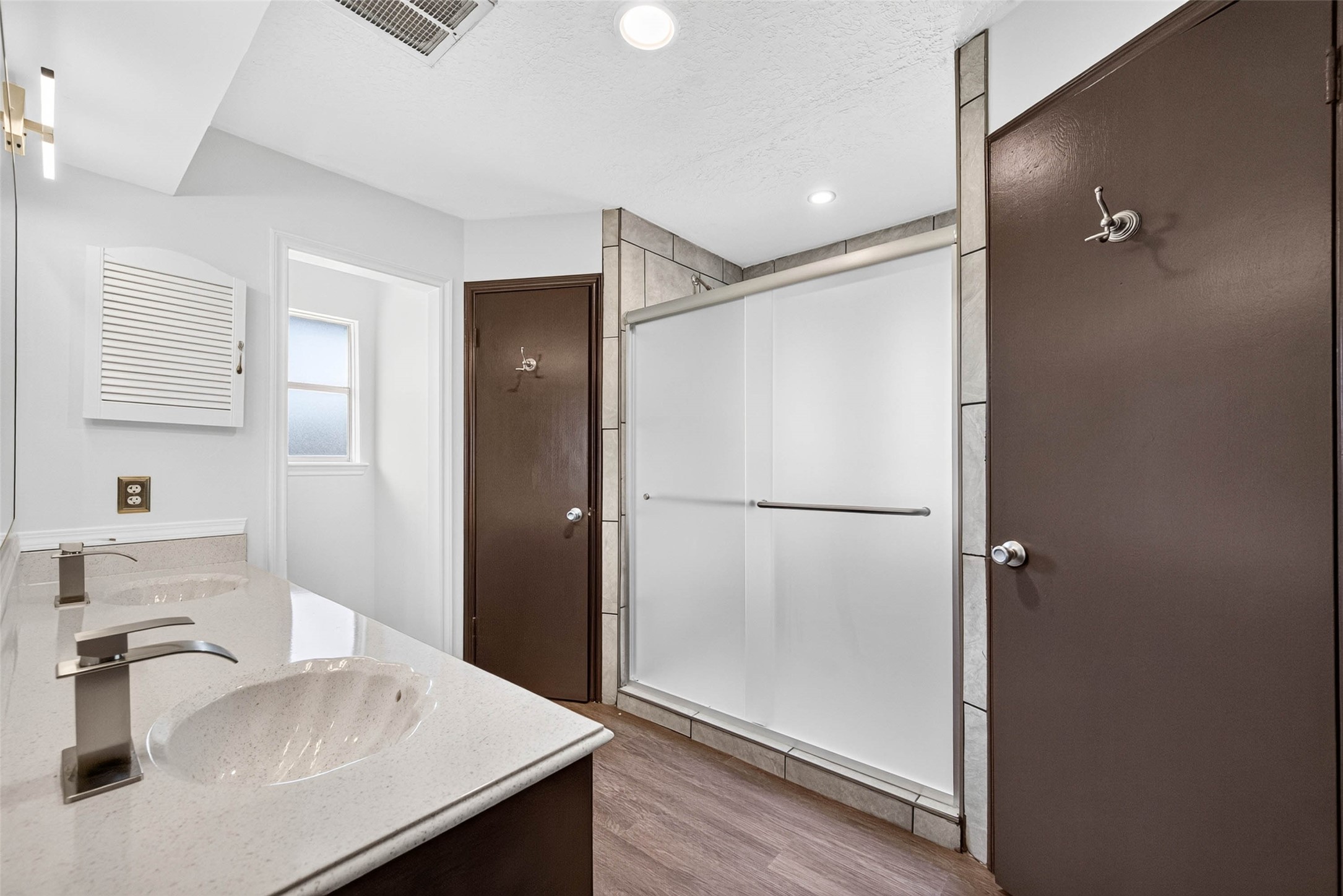 En-suite bathroom with a double vanity in dark wood, a speckled countertop, a frosted glass shower enclosure with tile surround, and wood-look flooring.