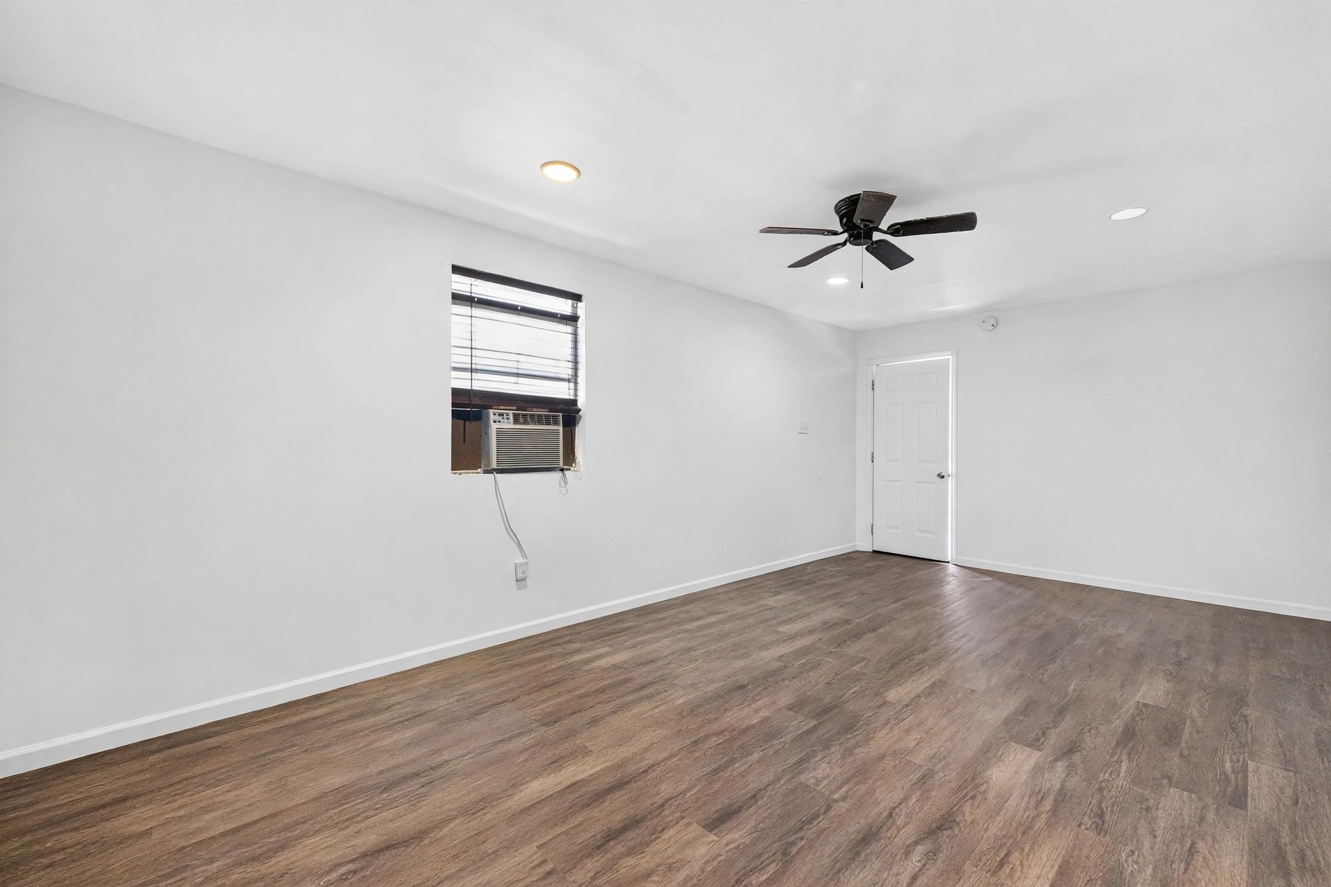 Another angle of the flexible interior room, with wood-look flooring, a dark ceiling fan, recessed lights, and a window air-conditioning unit.