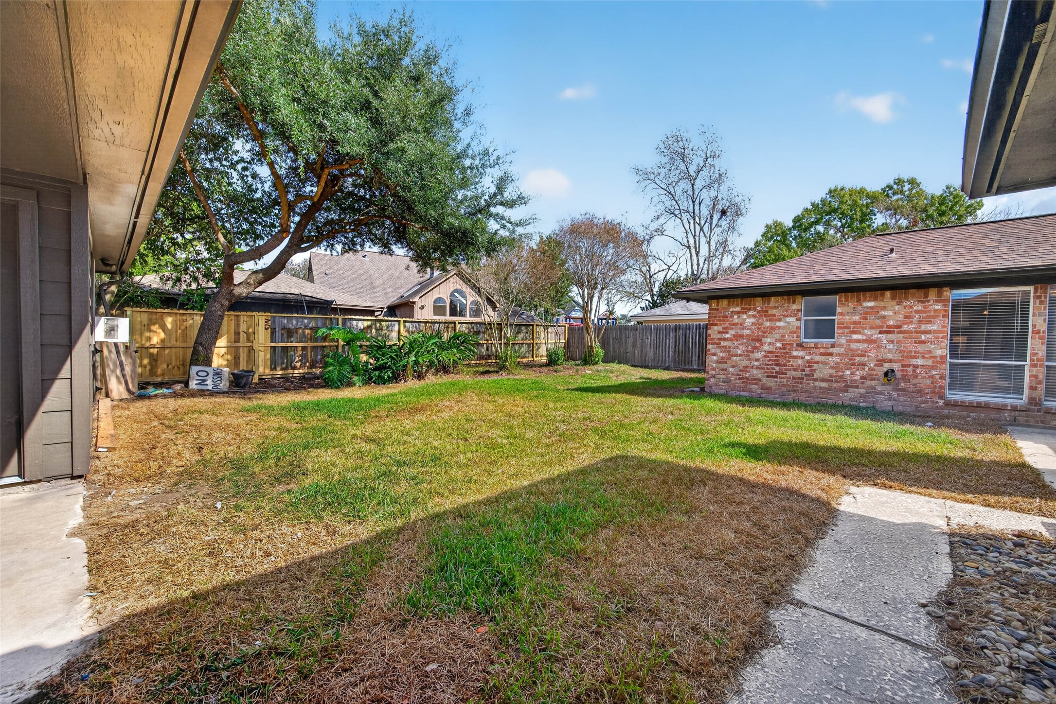 Wide backyard view across the lawn to the brick rear of the home, with a mature shade tree, wooden fencing, and a concrete walk.