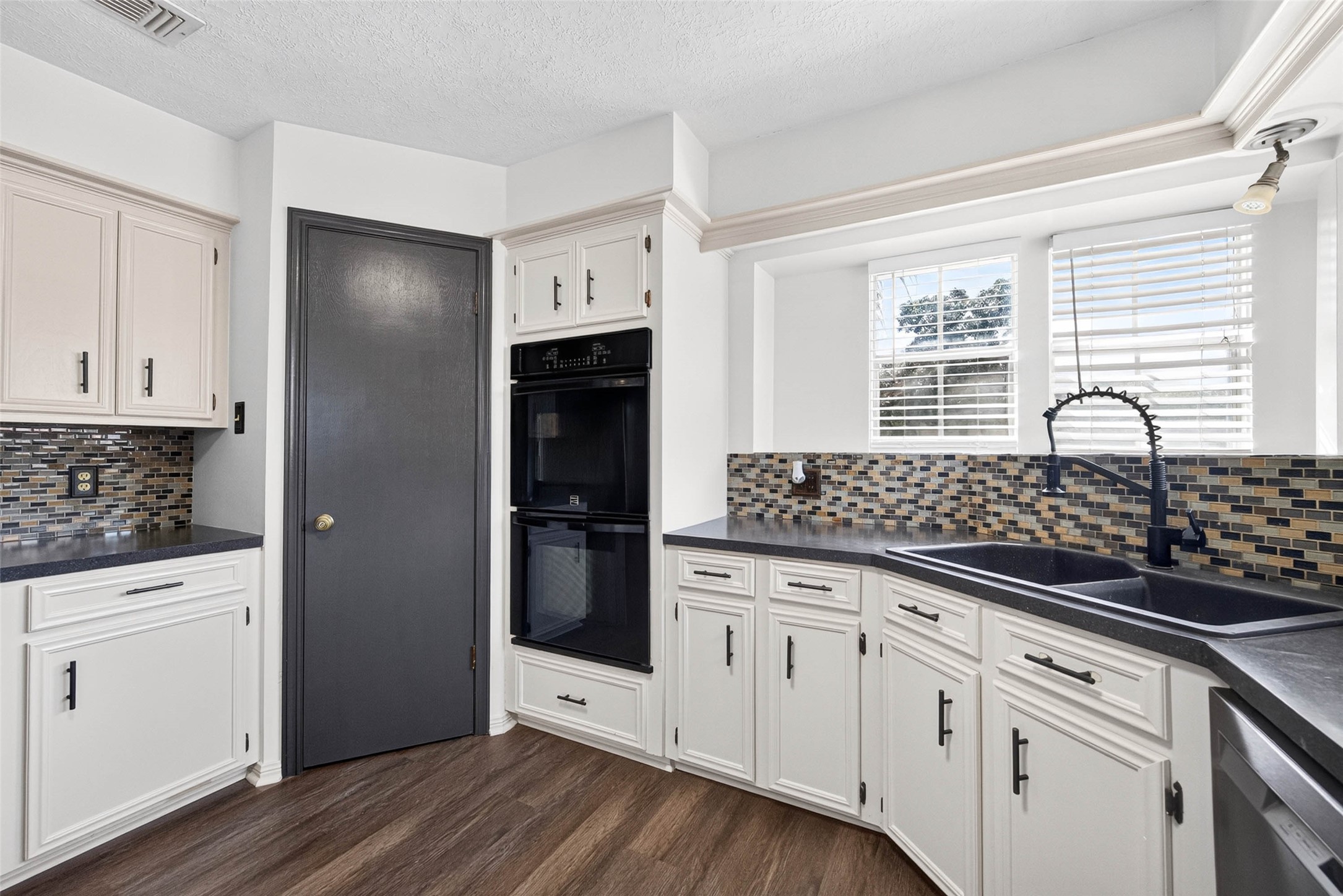 Kitchen view centered on the dark double-basin sink with a gooseneck faucet, set beneath windows with blinds and flanked by cabinets and the black double oven.