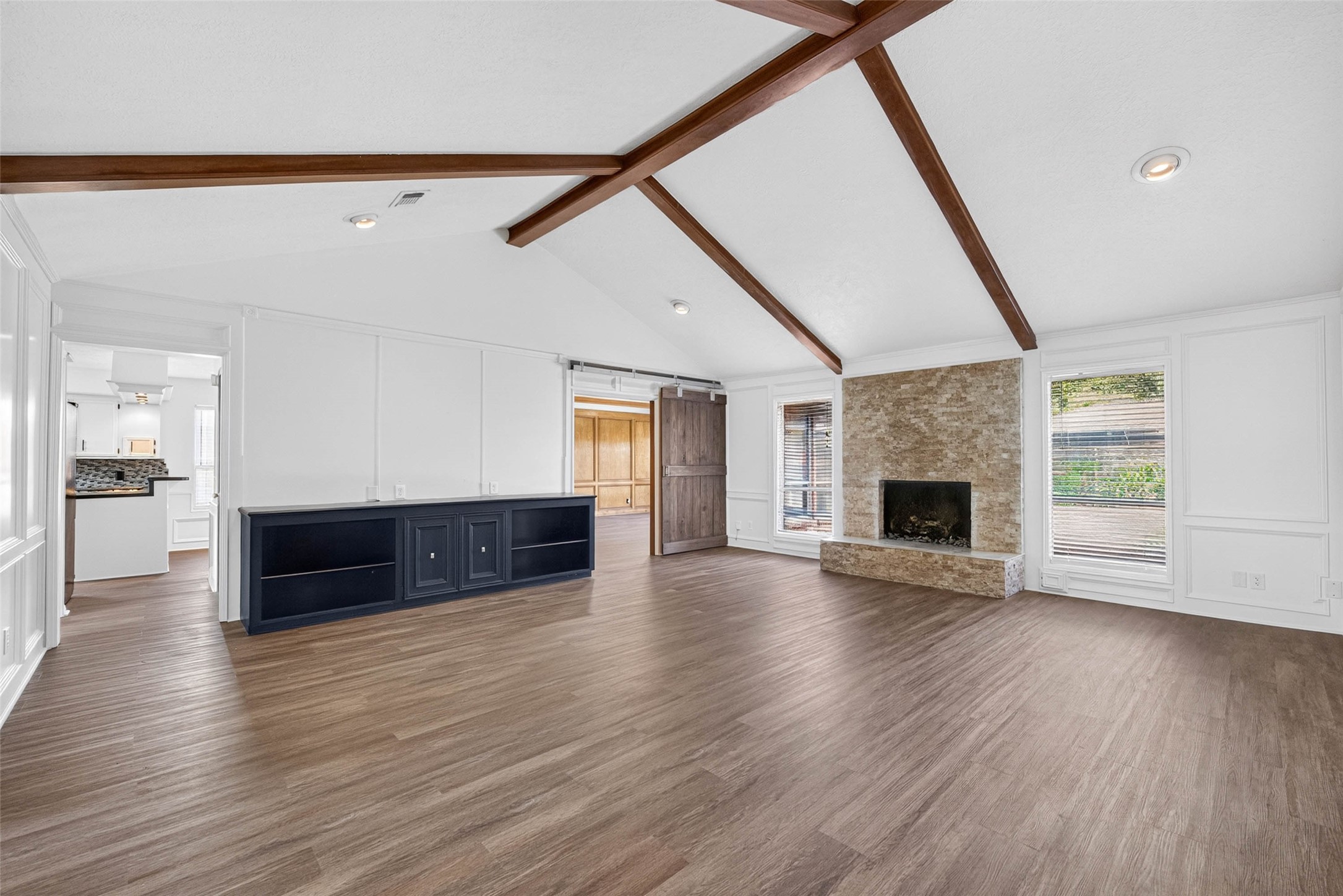 Living room under a vaulted ceiling with crossing dark wood beams, a stacked-stone fireplace, paneled walls, and a dark wood sliding barn door opening to the paneled room.