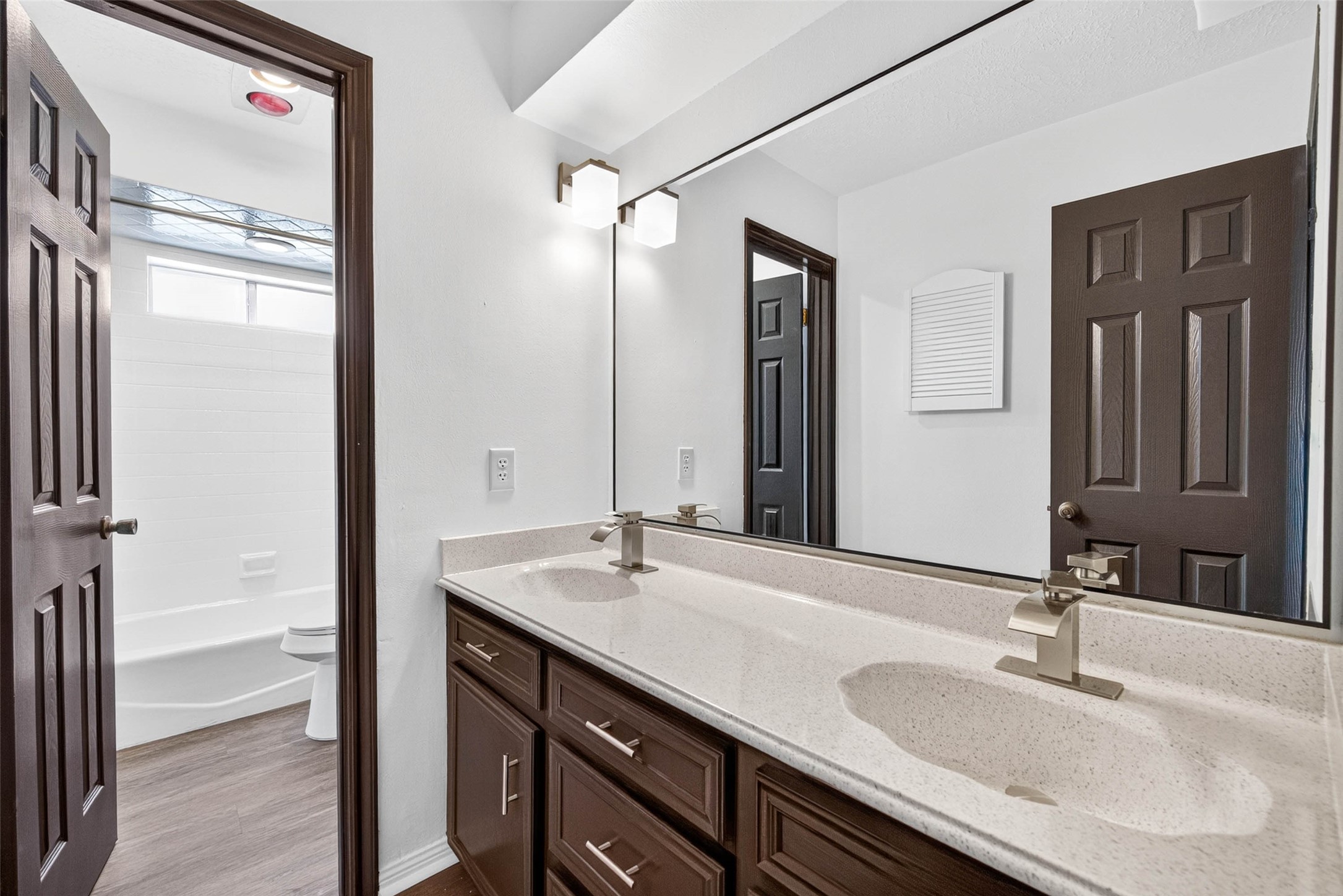 Bathroom with a long double vanity in dark wood, a speckled countertop, and a large mirror, with the tub and toilet room opening to the left.