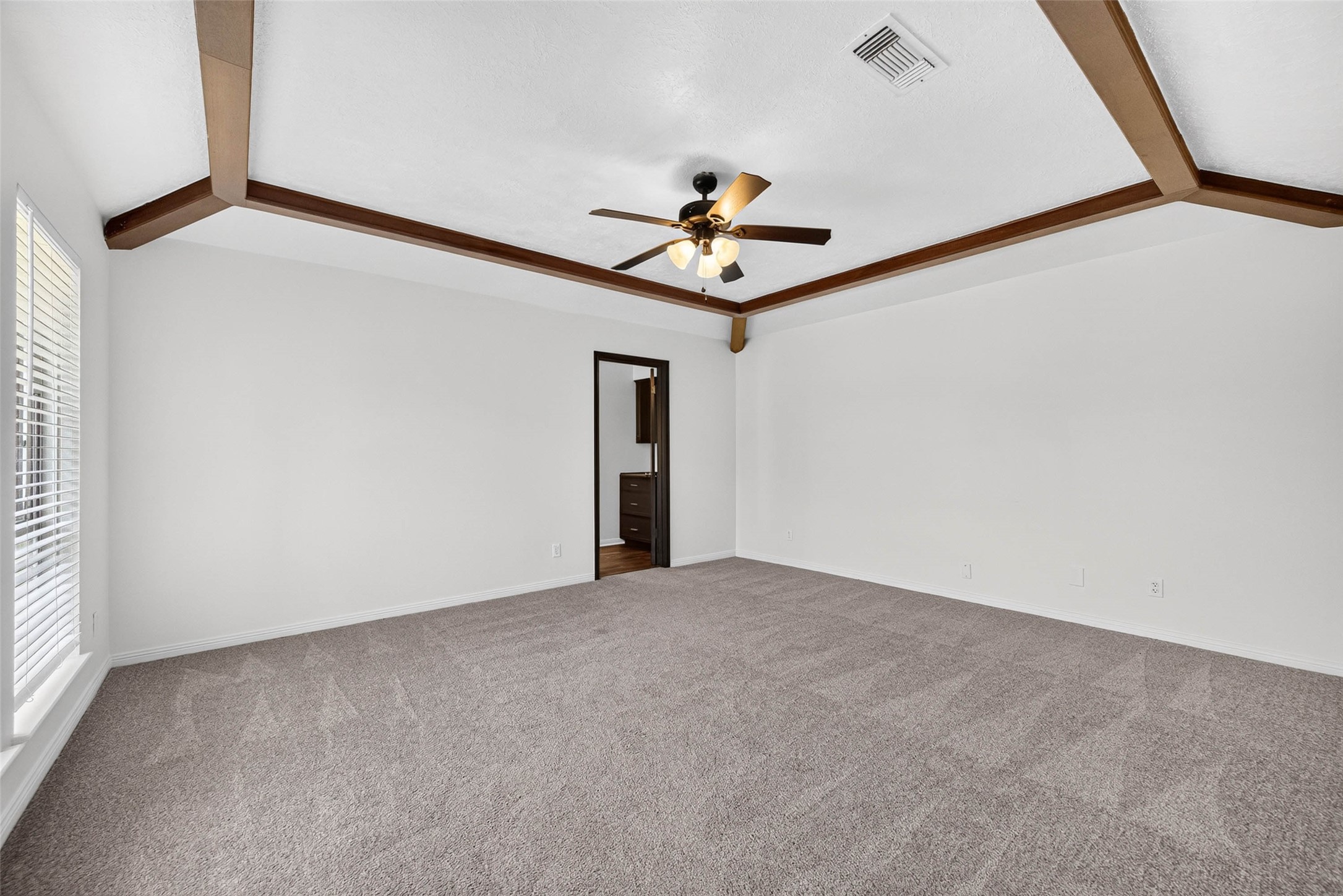 Large carpeted bedroom under a vaulted ceiling with dark wood beams and trim, a ceiling fan with lights, and a doorway to the en-suite bath.
