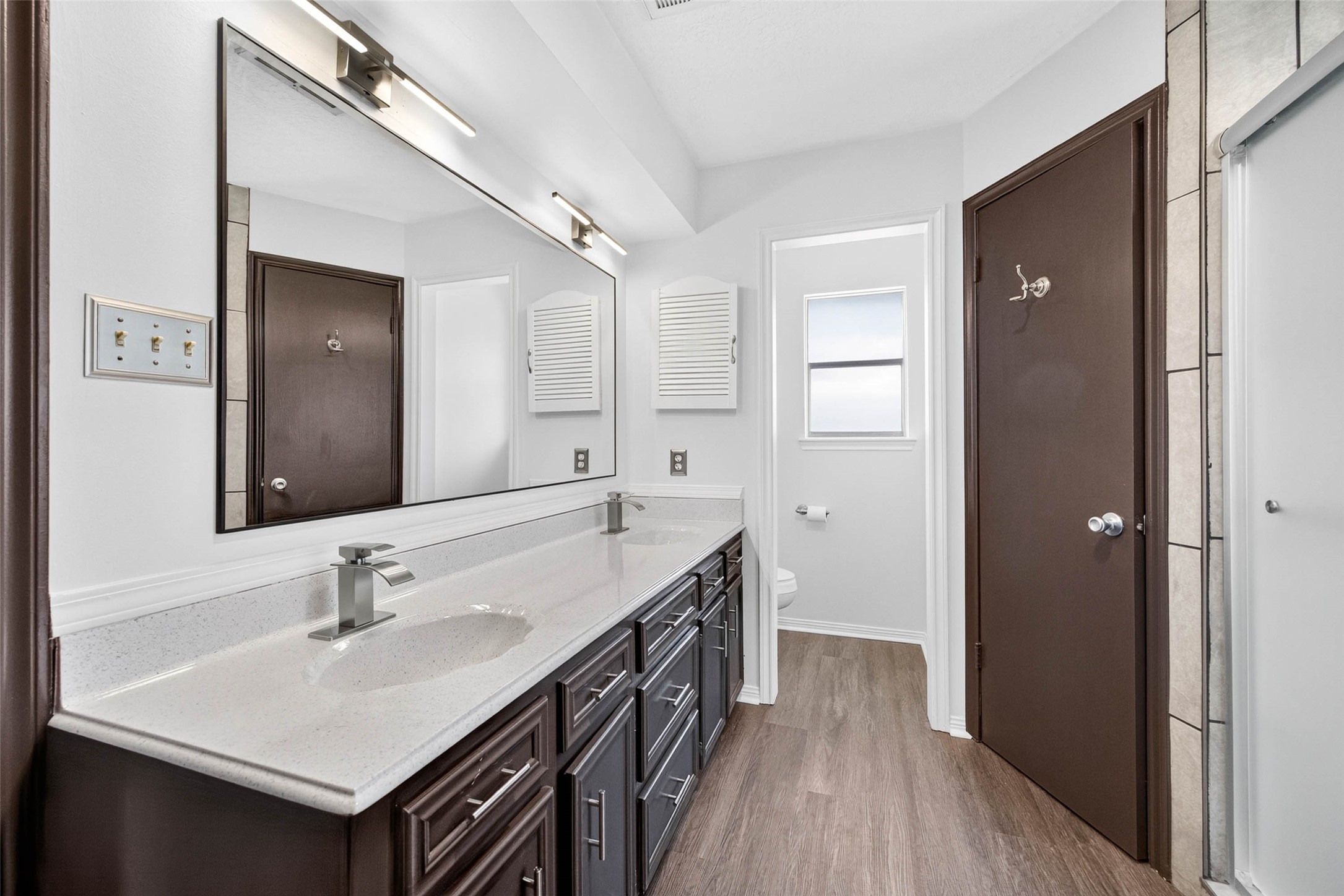 En-suite bathroom with a long double vanity, large mirror, modern lighting, and a separate toilet room with a window, all on wood-look flooring.