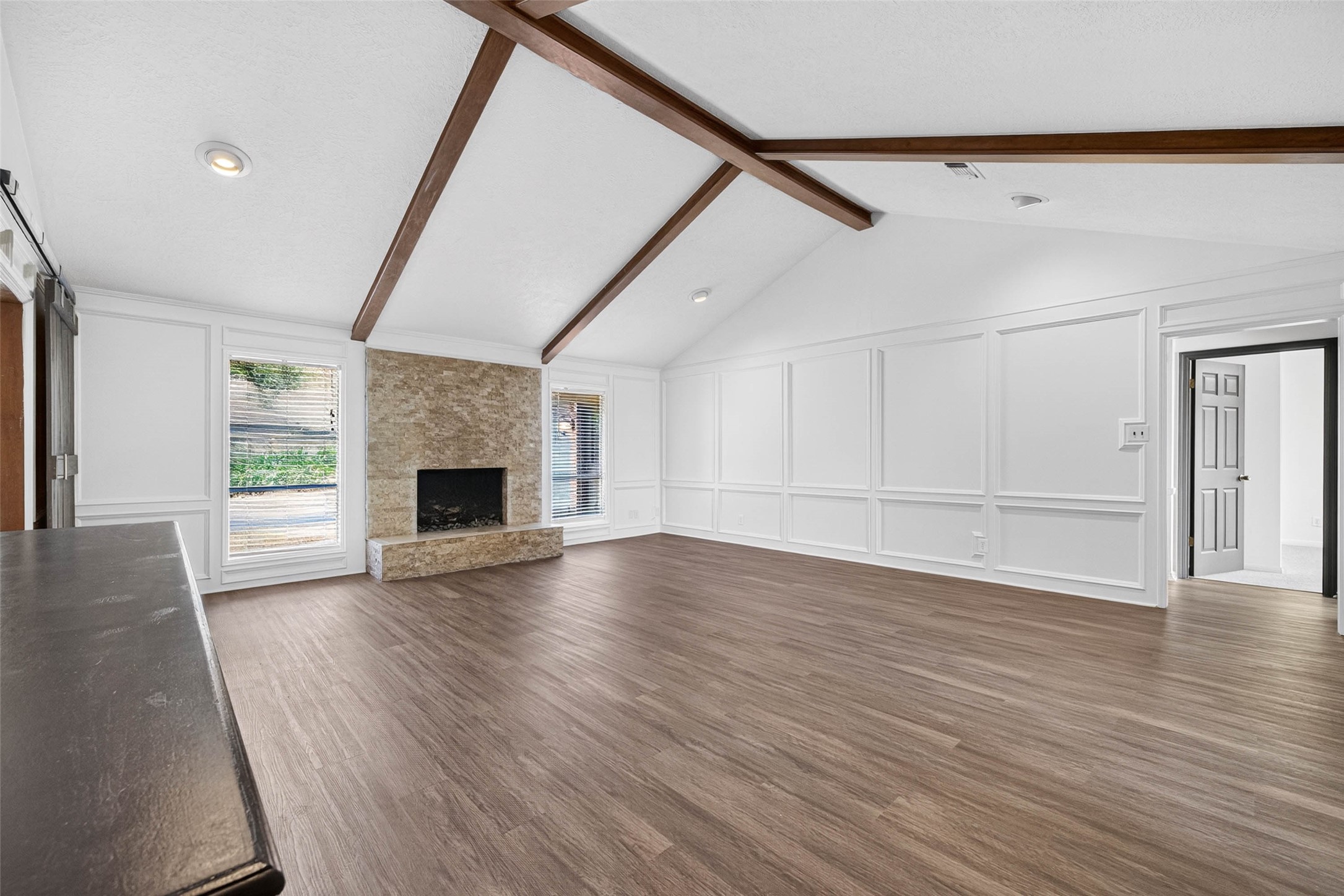 Living room angle toward the stacked-stone fireplace with raised hearth, flanked by windows with blinds, under crossing wood beams.