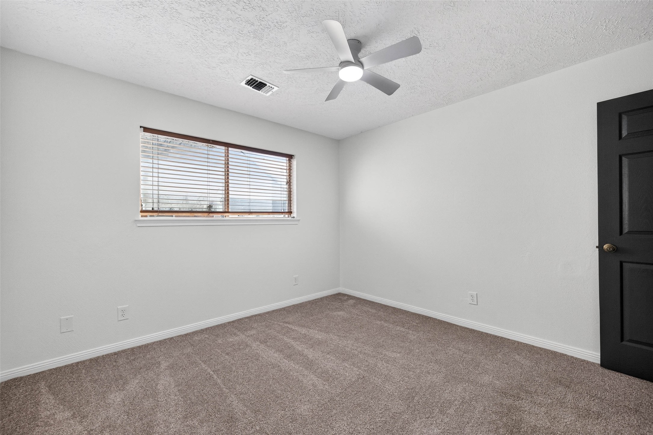 Carpeted bedroom with a wide window with blinds, a ceiling fan with light, and a dark interior door.