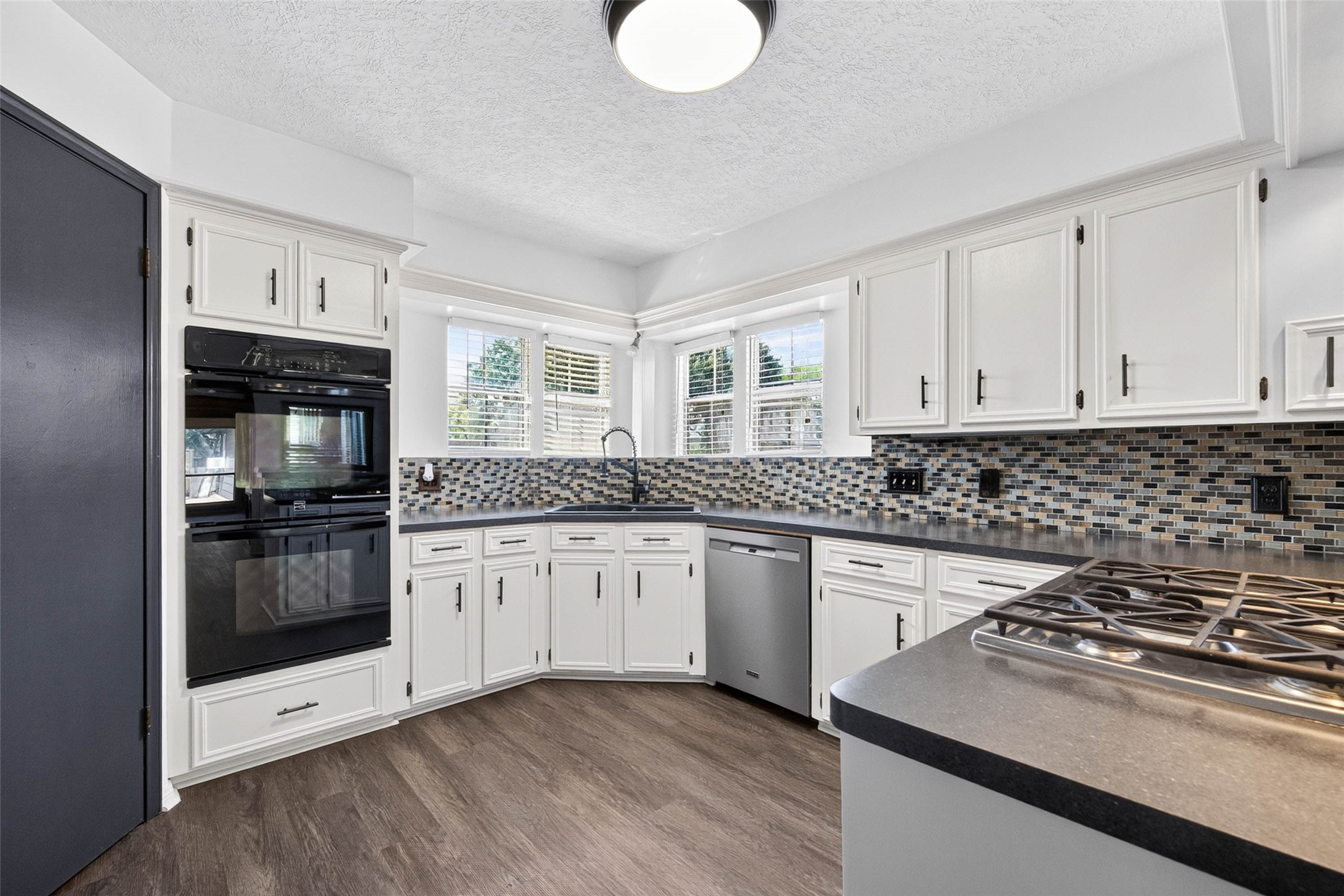 Kitchen angle showing cabinets, a mosaic tile backsplash, black double wall oven, corner sink beneath windows, and a gas cooktop on the peninsula.