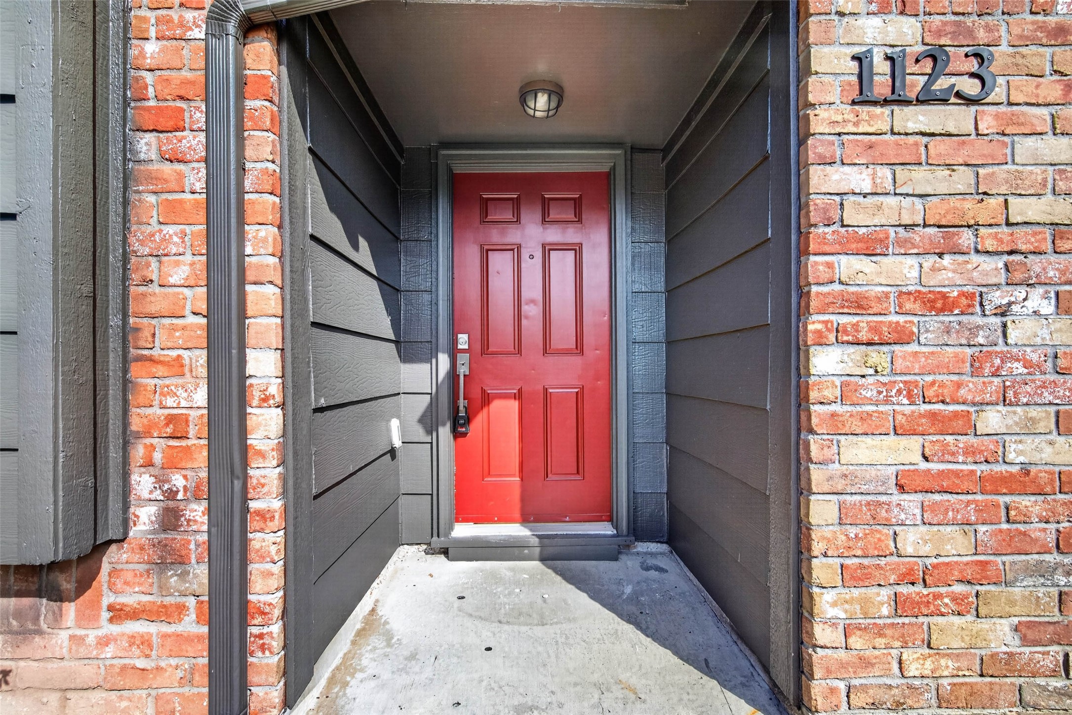 Recessed front entry with a red paneled door, dark gray siding walls, and the house number 1123 in dark metal on the brick wall.