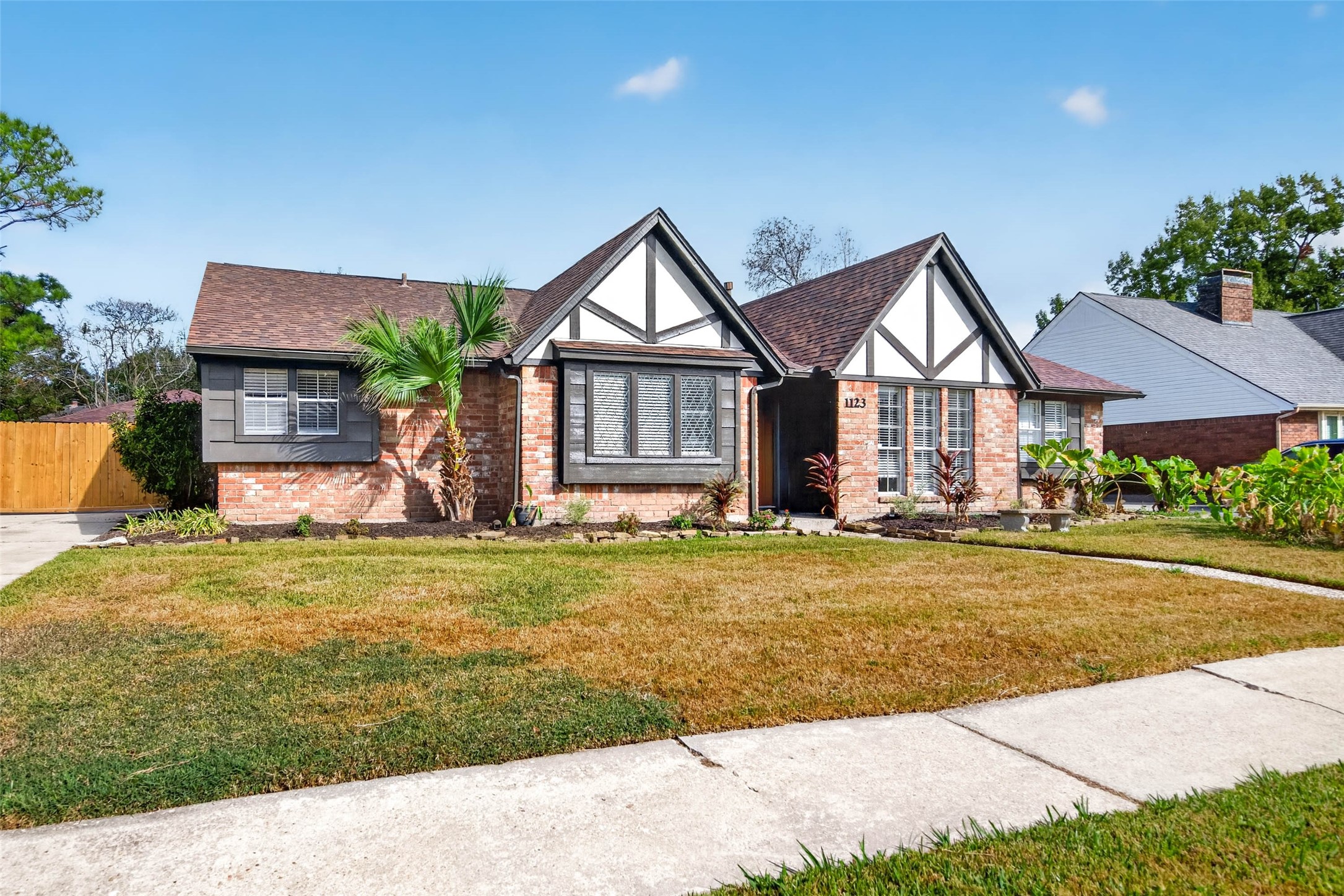 Front view from the sidewalk across the lawn, with the white gables and dark timber trim, bay window, palm, and wooden fence at the side.