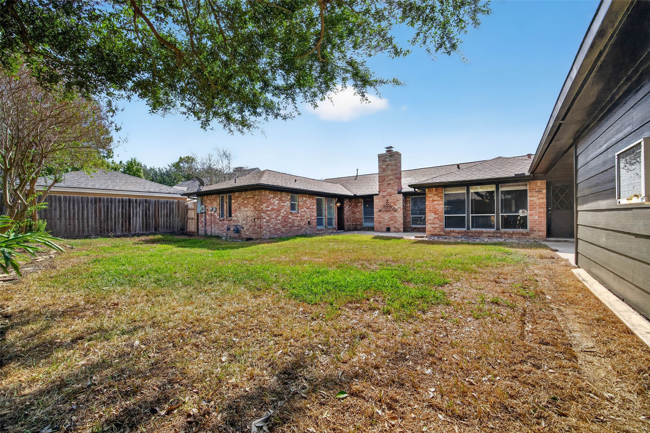 Wide backyard view of the brick rear of the home with its chimney and large windows, framed by lawn, fencing, and mature trees.