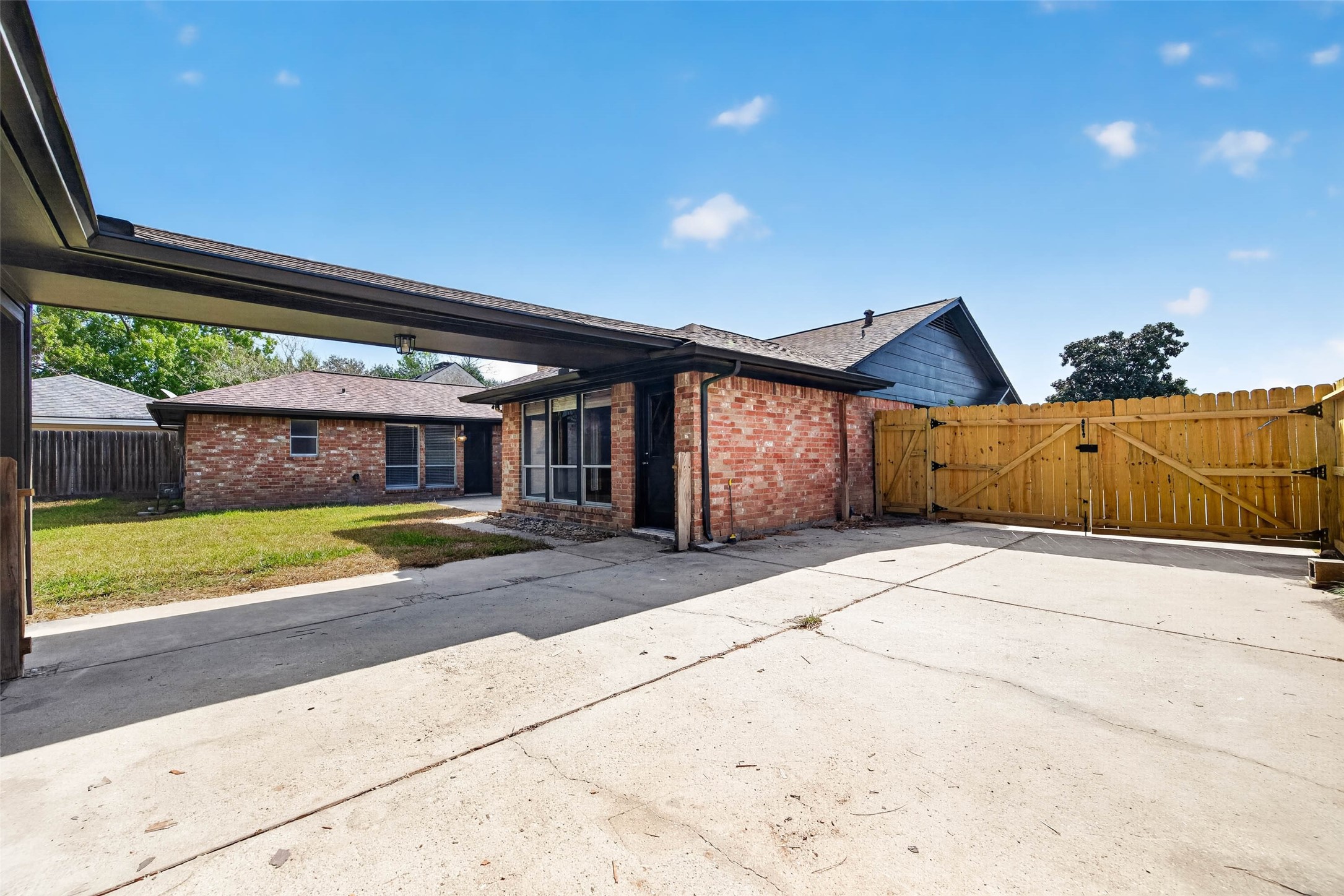 Covered area and concrete driveway beside the brick house, with a wooden double gate opening to the lawn.