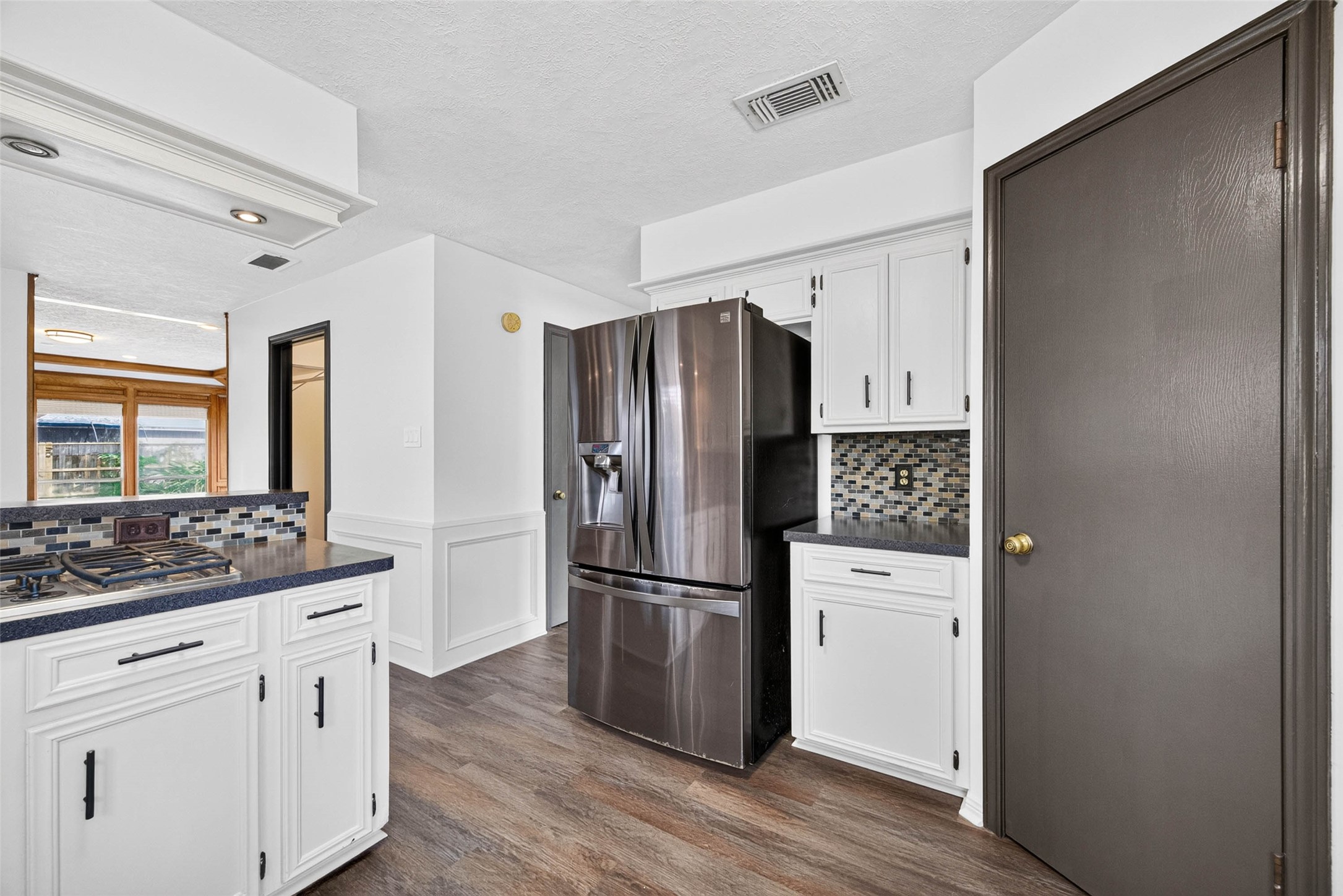 Kitchen angle with a stainless French-door refrigerator, cabinets, mosaic tile backsplash, and a gas cooktop, with the paneled living room visible beyond.