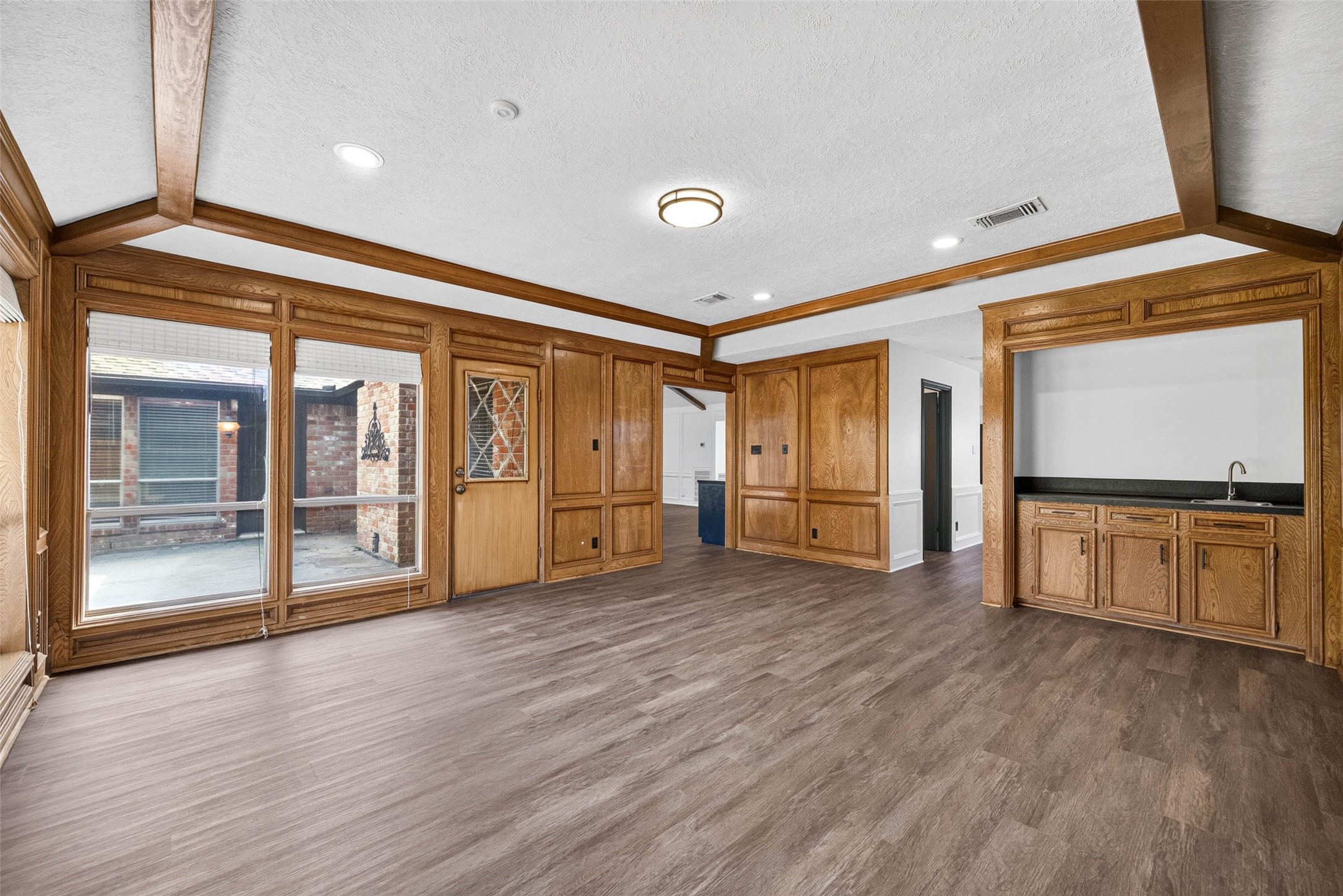 Paneled living room view toward the bar, with glass-paneled wood doors, wide windows to the patio, and an open doorway to the kitchen.