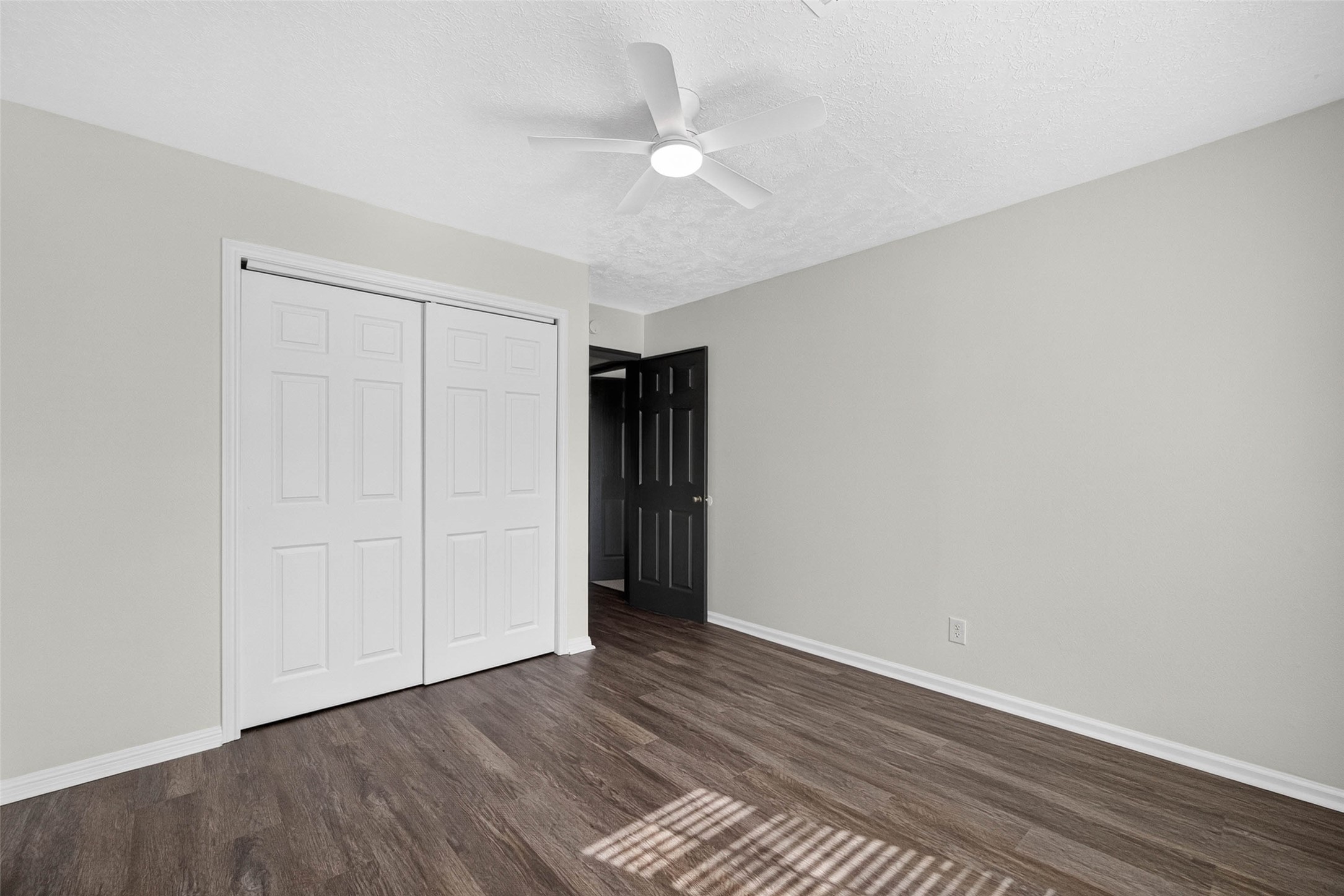 Bedroom with wood-look flooring, sliding closet doors, a dark interior door, and sunlight striping the floor through blinds.