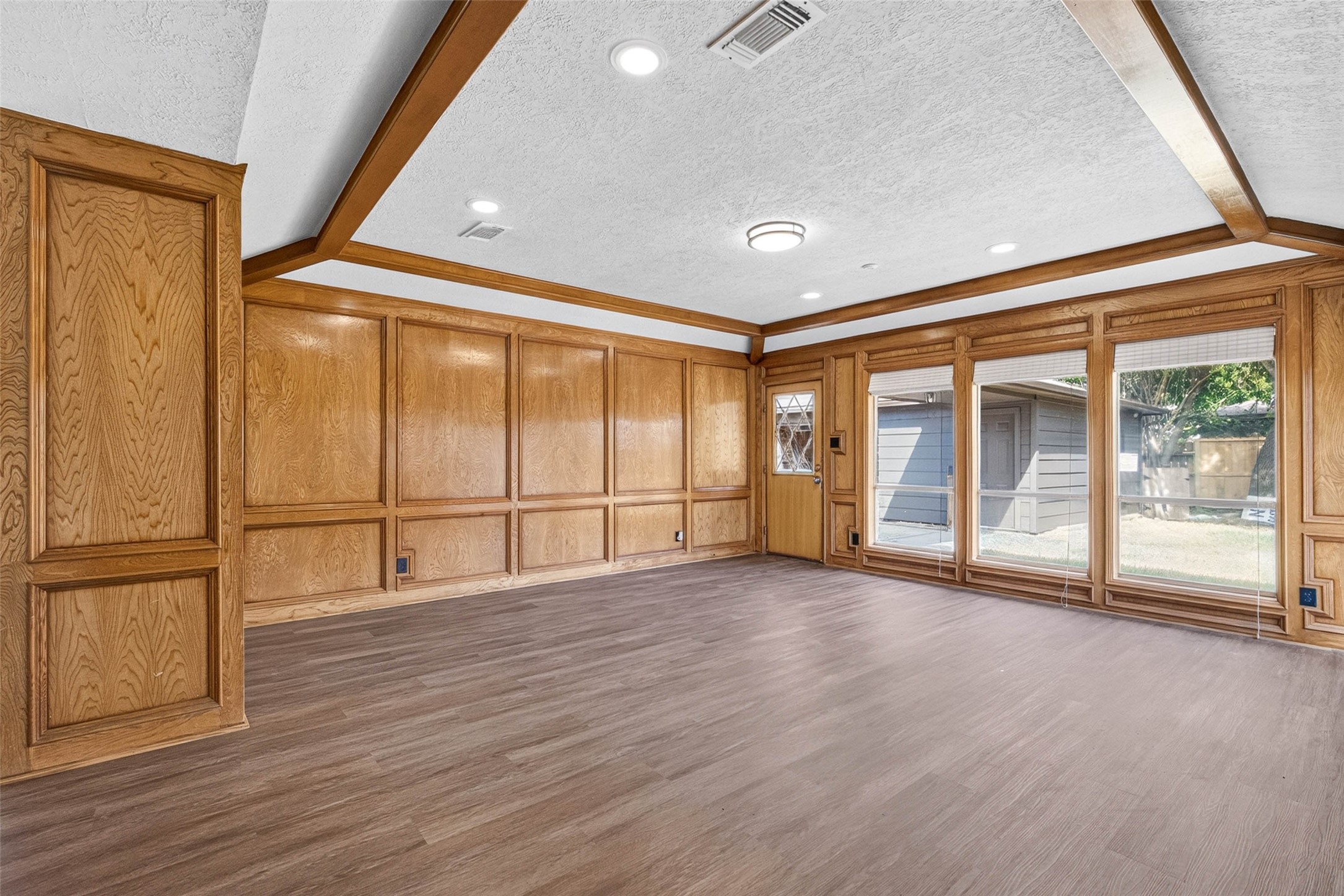 Wood-paneled walls with raised panel detailing, exposed wood beams, and large windows overlooking the yard set the tone for this open living room on wood-look flooring.