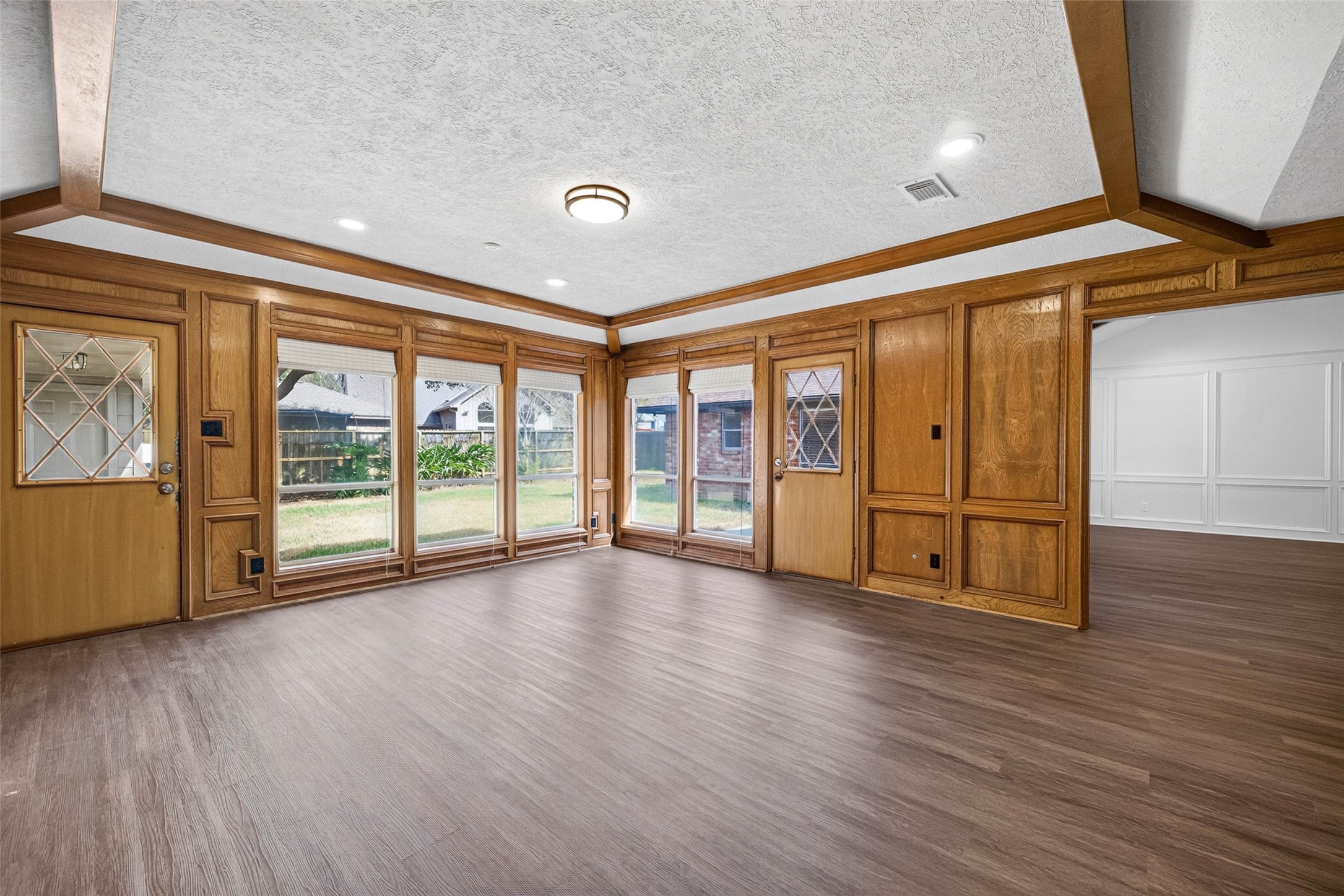 Corner view of the paneled living room, where a full wall of windows frames the backyard and two glass-paneled wood doors open to the patio.