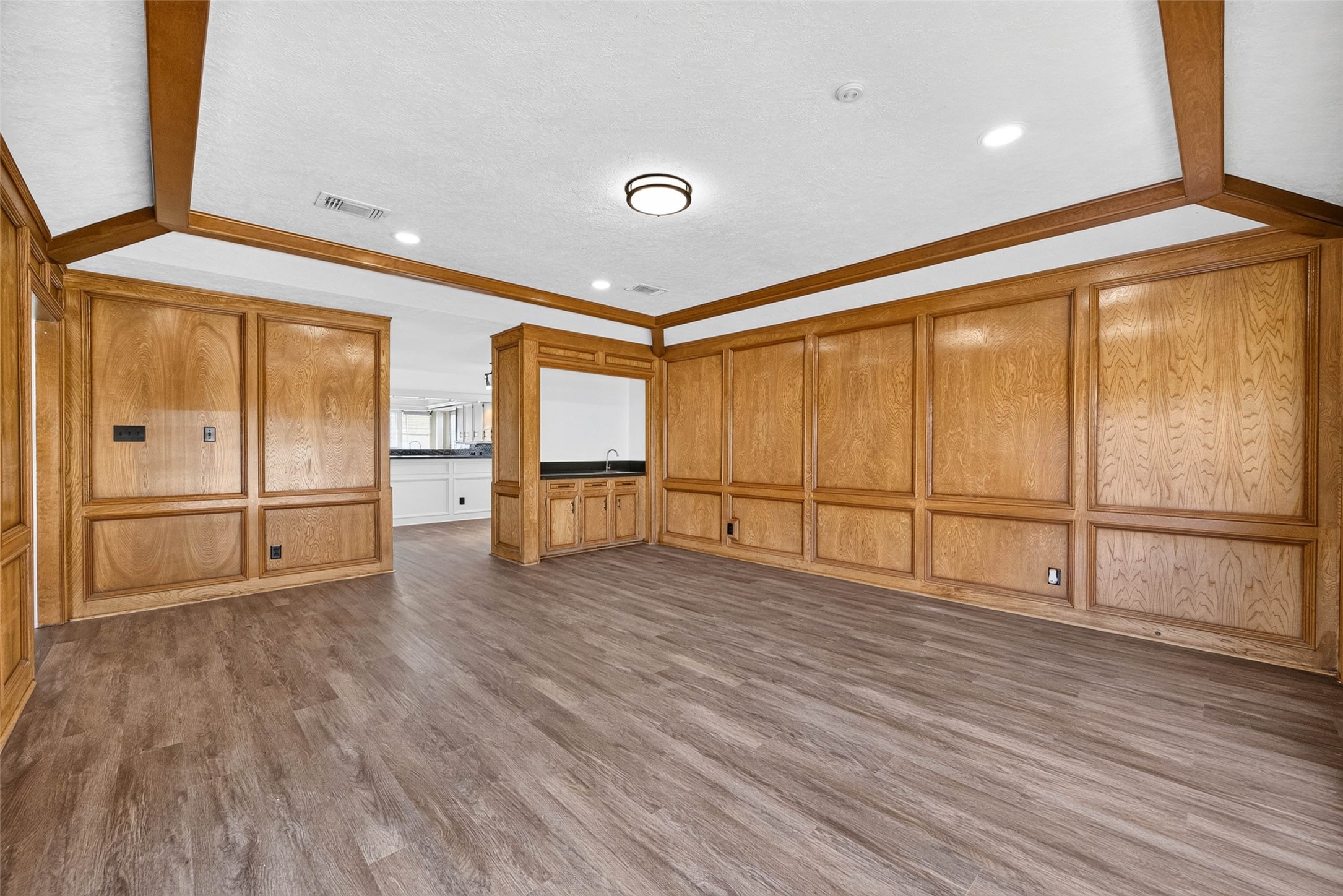 nother angle of the paneled living room, where the wood beams and wood-look floors carry through to a bar nook and the kitchen beyond.