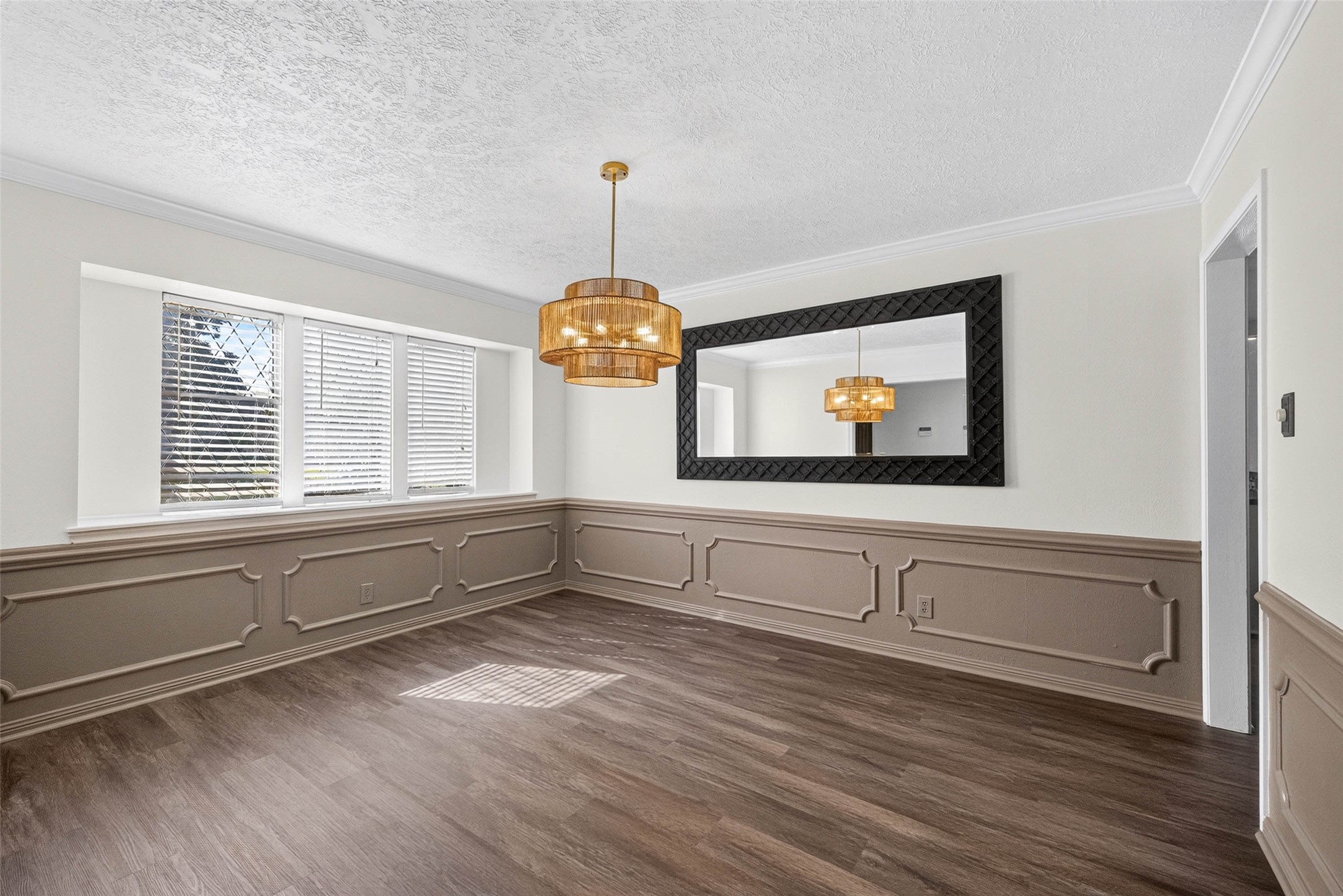 Another angle of the front room, with wainscoted walls, three windows with blinds, a tiered pendant light, and a doorway to the next room.