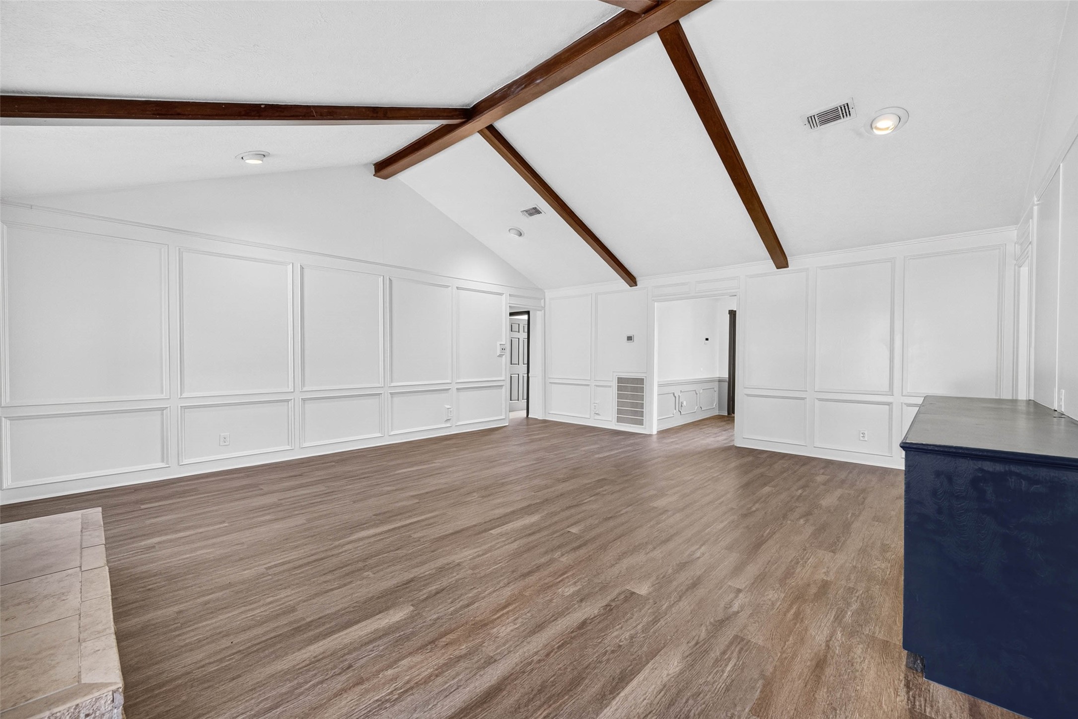 Living room view across wood-look flooring to full-wall panel molding, crossing beams, and the stone hearth, with a doorway to the front room.