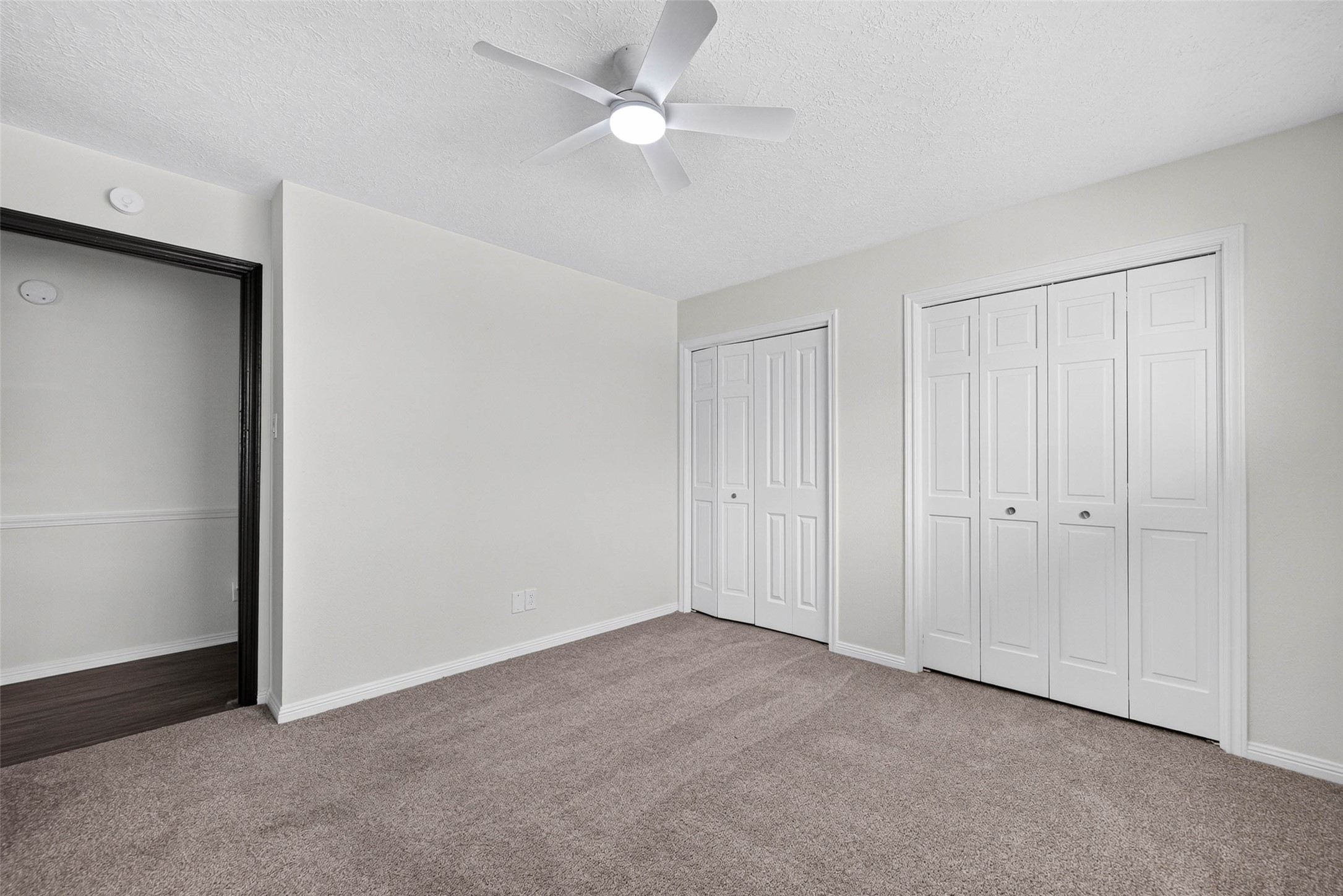 Carpeted bedroom with two sets of closet doors, a ceiling fan with light, and a doorway opening to a wood-look floored room.