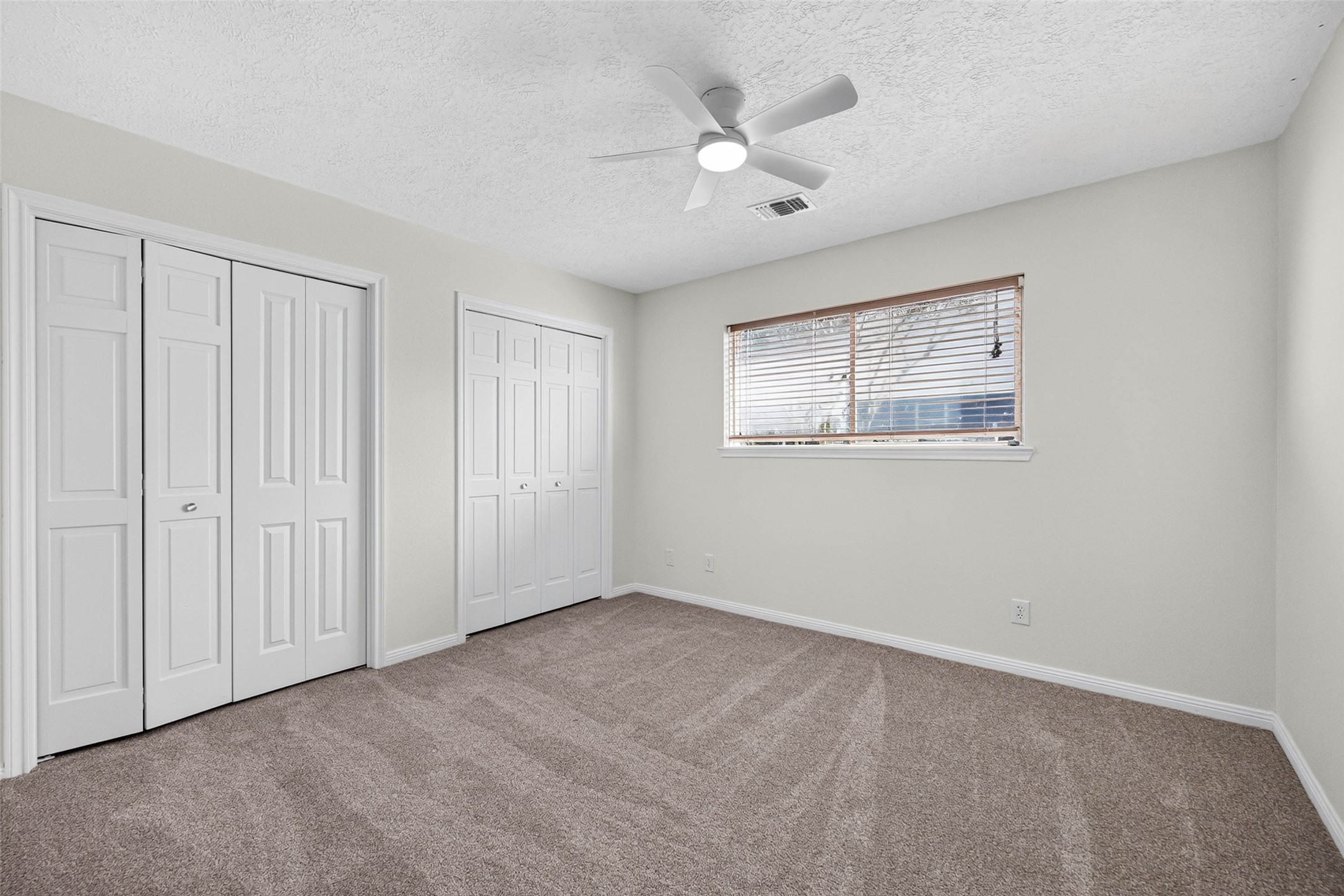 Carpeted bedroom with two closet door sets and a wide window with blinds, offering flexible furniture placement.