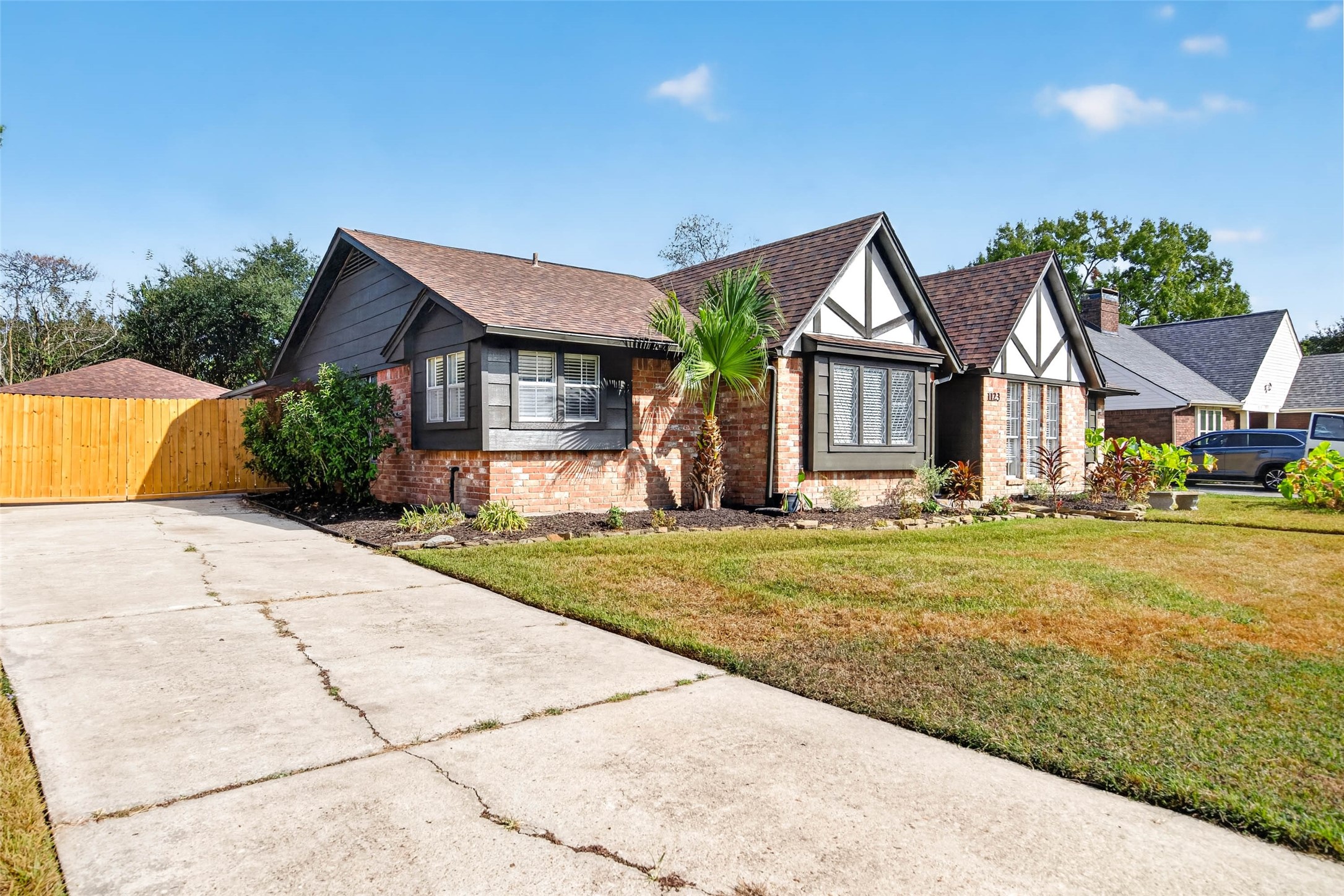 Angled front view from the concrete driveway, showing the brick and dark siding exterior, brown shingle roof, wooden side fence, and landscaped bed with stone edging.