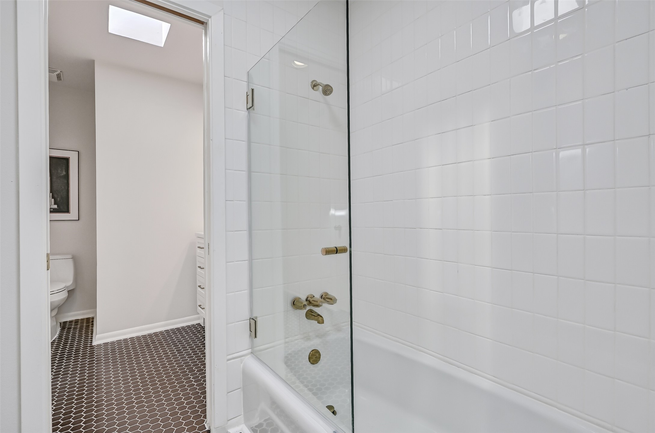 The shared bathing area features a tub/shower combination with white tiled walls, a frameless glass panel & gold-tone plumbing fixtures. Black hexagonal tile flooring continues throughout, while a separate toilet compartment and individual vanity areas provide added functionality for both bedrooms.