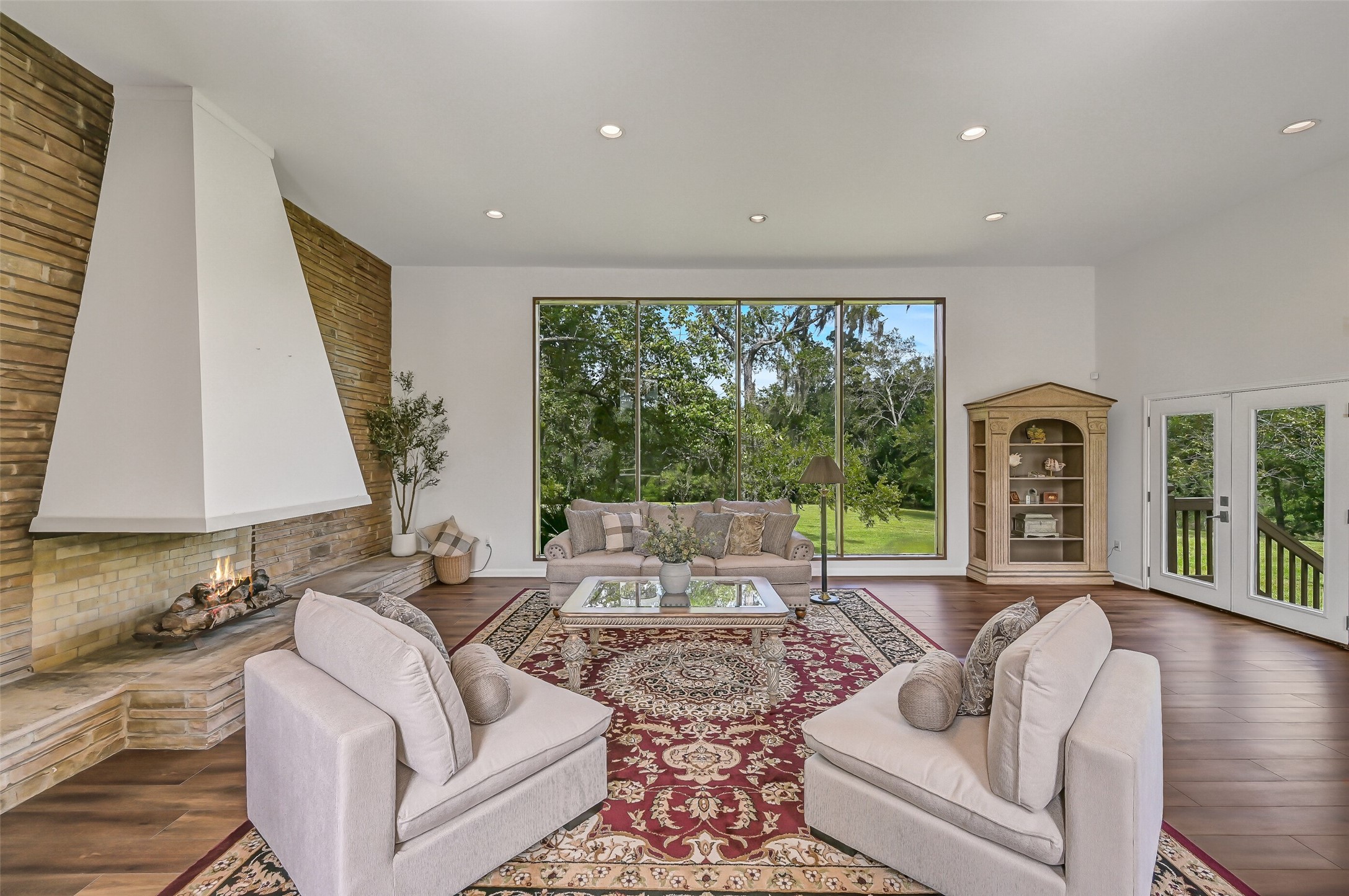 An impressive wall of picture windows brings the outdoors into the spacious living room, offering sweeping views across the backyard and mature trees. The room's generous proportions provide flexibility for multiple seating arrangements, while the distinctive fireplace creates a striking focal point.